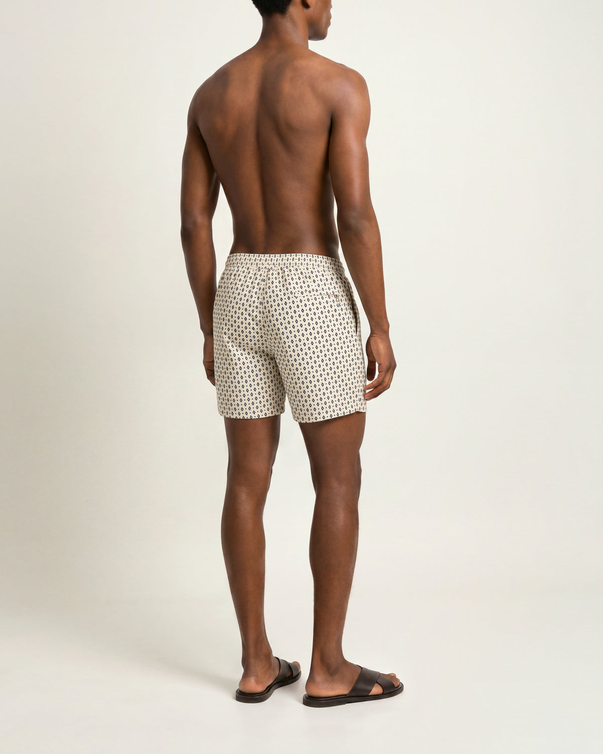 Back view of a man wearing beige patterned drawcord swim shorts and brown sandals on a plain background
