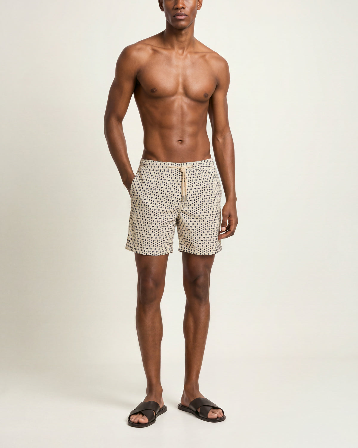 Man wearing beige patterned drawcord swim shorts and brown sandals on a plain background