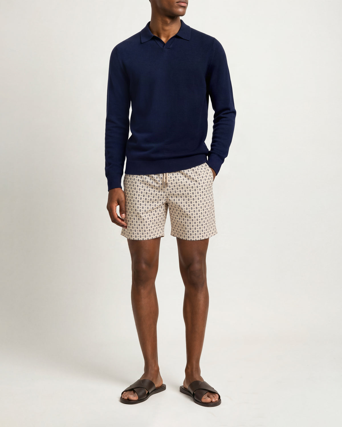 A man modeling a navy blue long-sleeve polo sweater paired with beige shorts featuring a small black geometric pattern, styled with dark brown slide sandals against a plain light gray studio background.