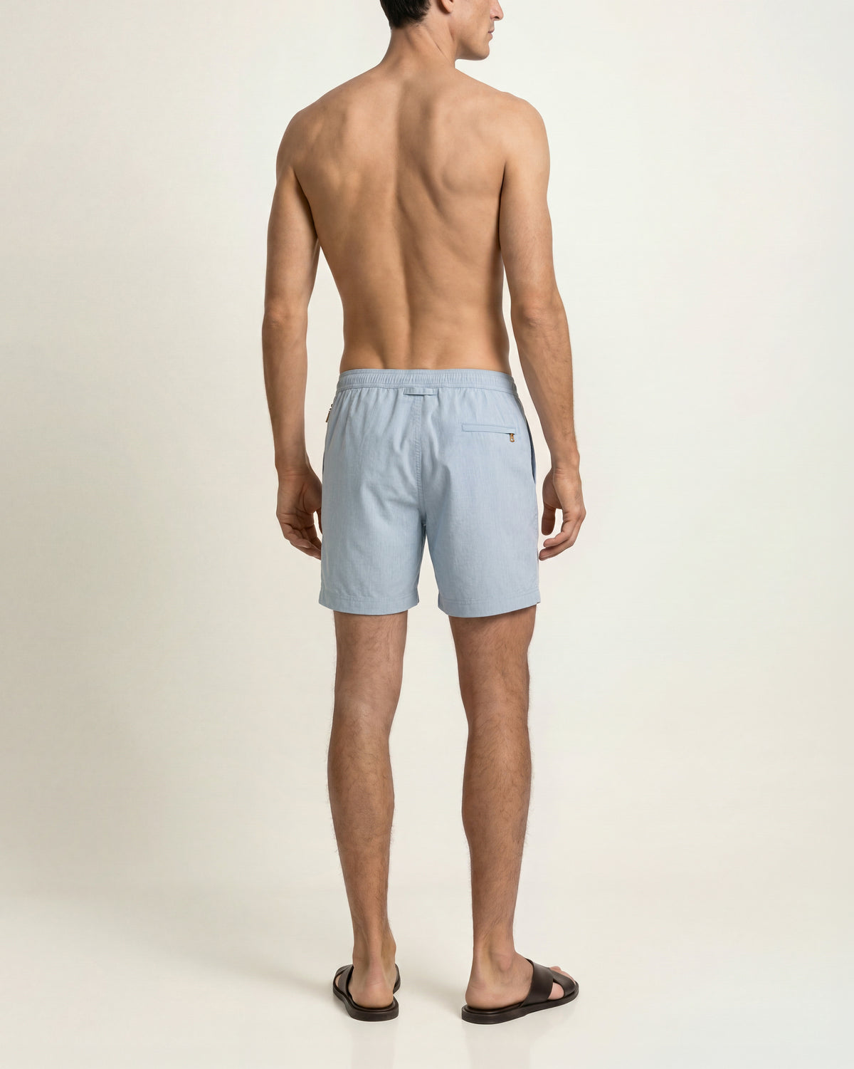 Back view of a man wearing light blue drawcord swim sorts and brown sandals on  a plain background