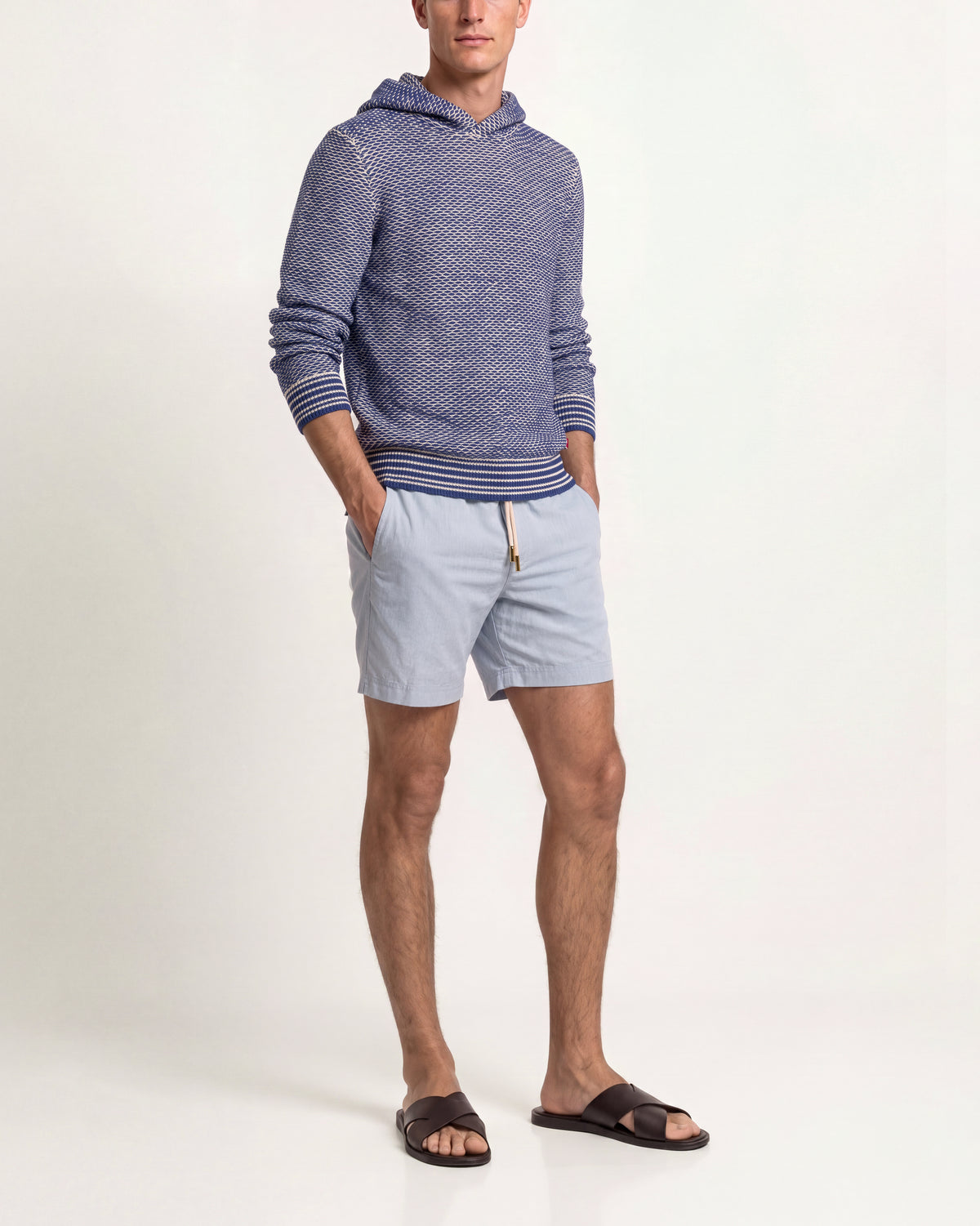 A man modeling a blue and white textured knit hoodie with striped ribbed cuffs and hem, paired with light blue drawstring shorts and dark brown slide sandals, against a plain white studio background.