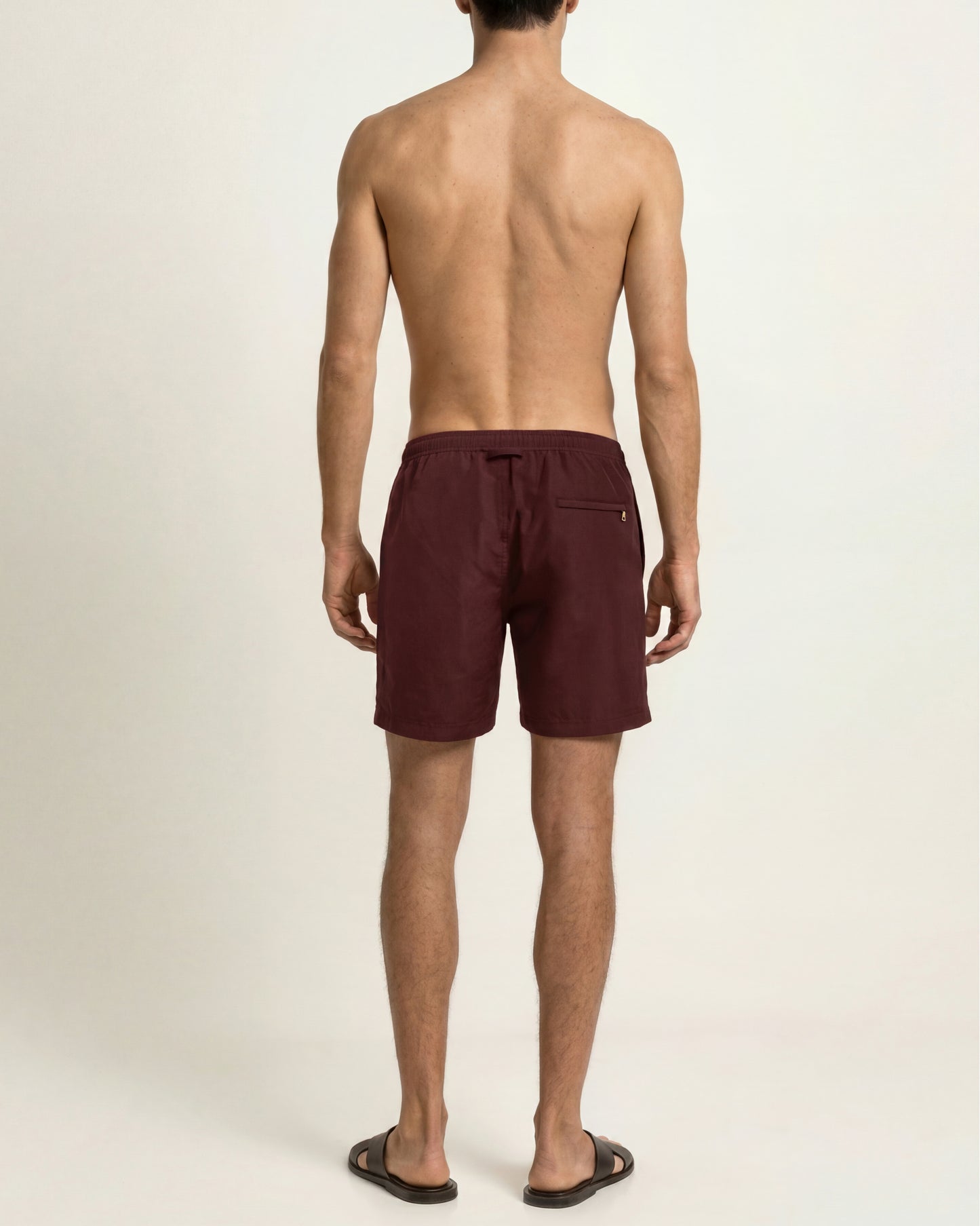 A man shown from the back wearing burgundy swim trunks and black sandals against a plain off-white studio background.