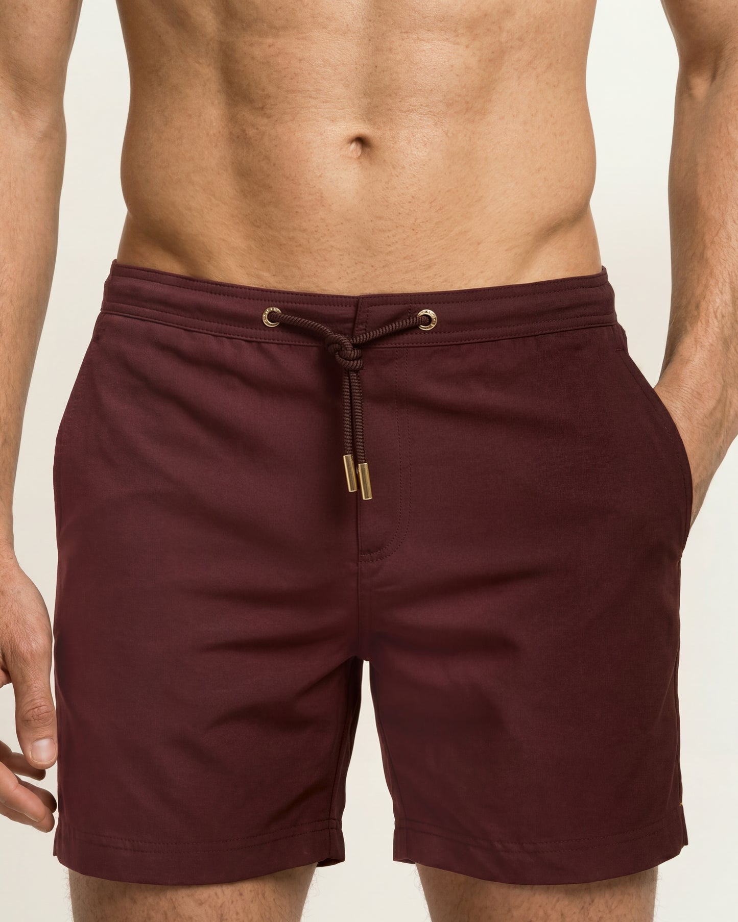 A close-up of a man wearing burgundy swim trunks with a drawstring waist and gold-tone hardware, cropped from the chest down against a light neutral background.