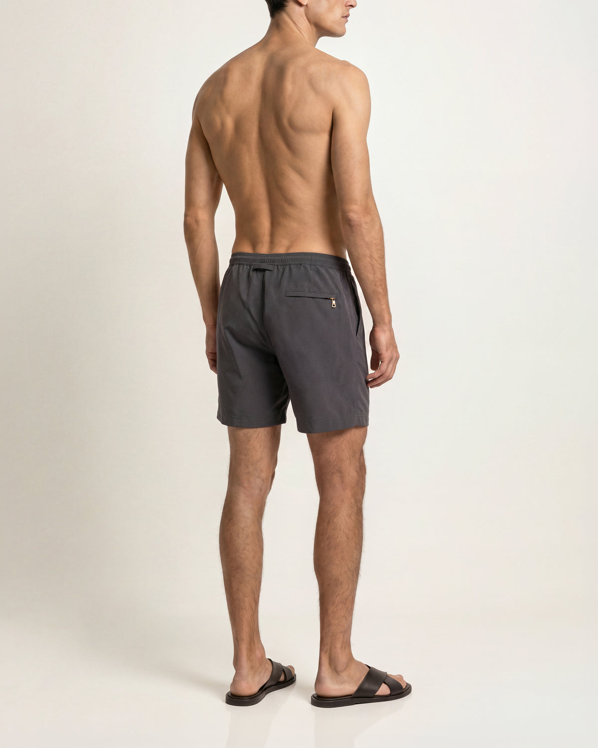 Man wearing grey swim shorts with brown sandals on a plain background