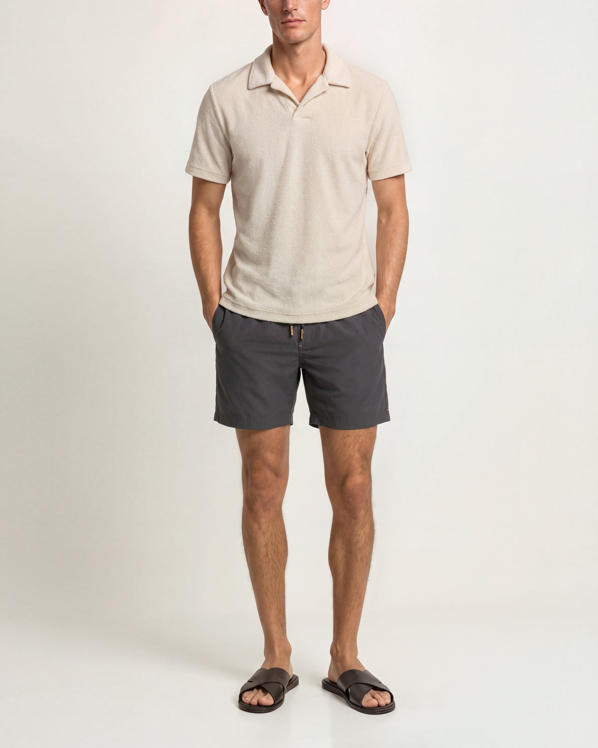 A man modeling a light beige short-sleeve polo shirt and dark gray shorts, paired with black slide sandals, against a plain off-white studio background.