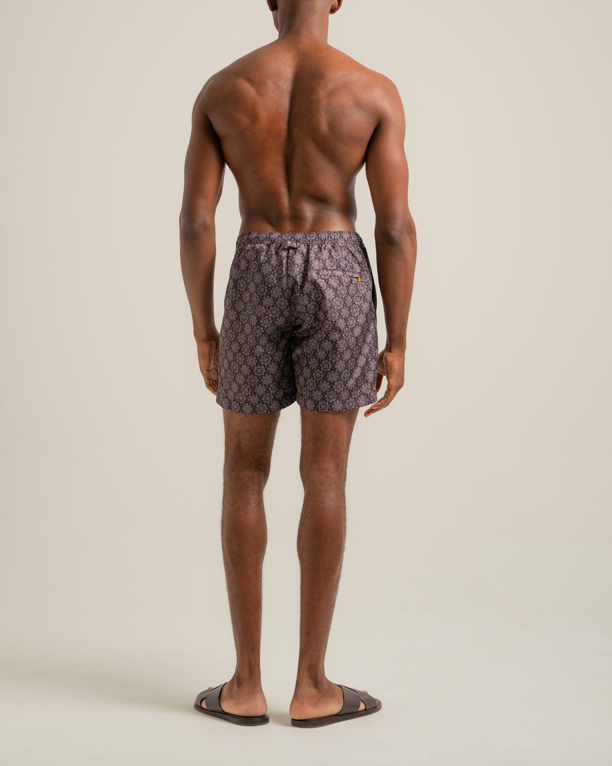 A man viewed from behind wearing patterned swim trunks with a small gold logo on the left side, standing against a plain light beige studio background.