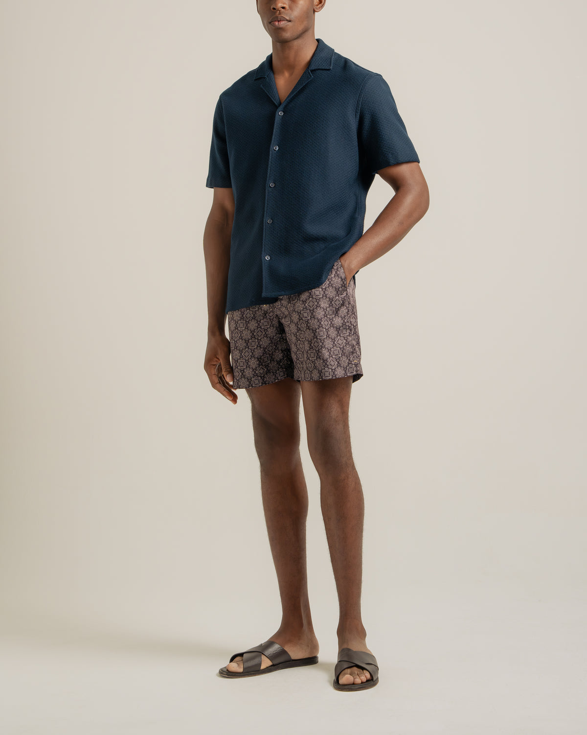 A man modeling a dark navy blue short-sleeve button-up shirt and patterned shorts with brown slide sandals, standing against a solid light beige studio background.