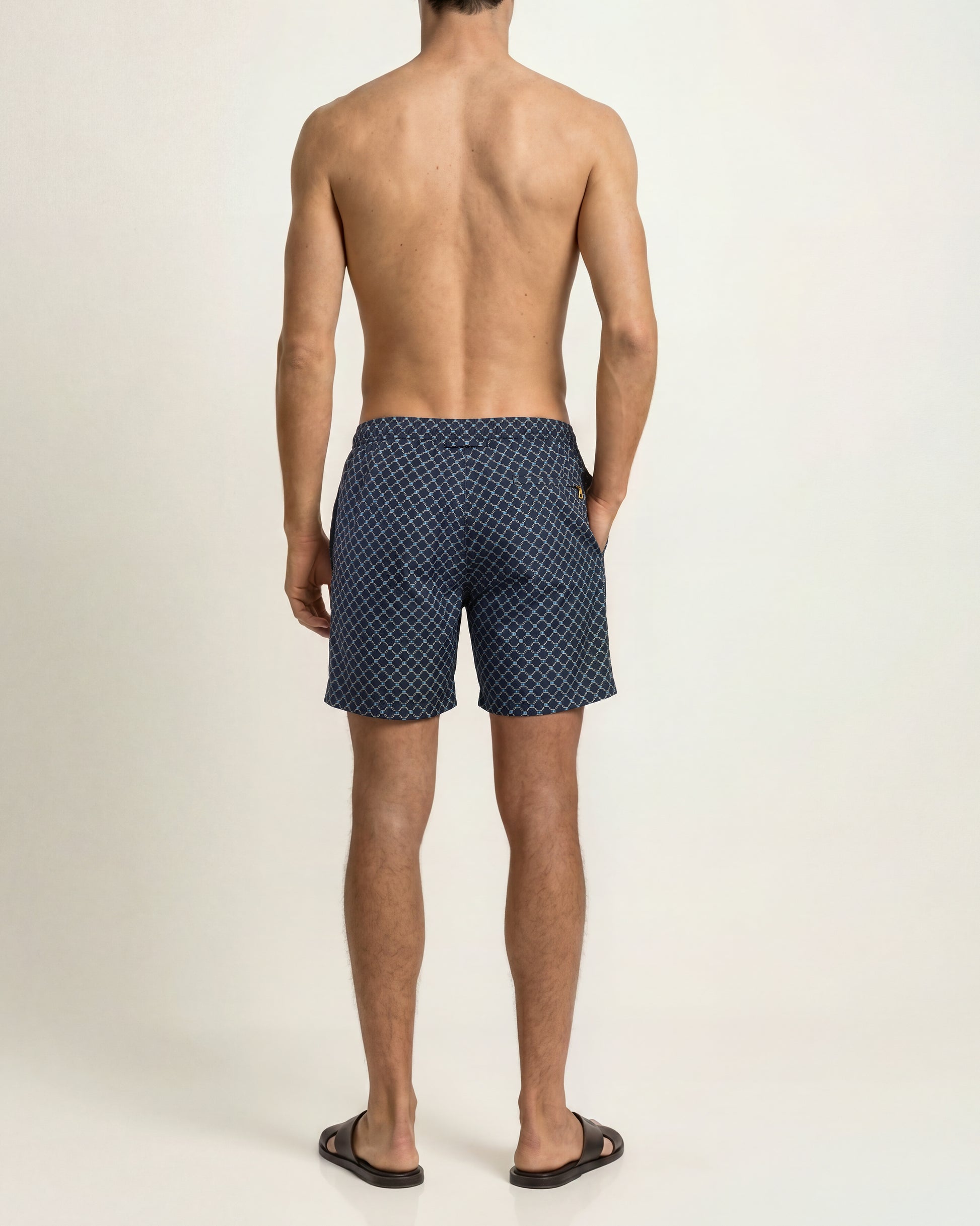 A man viewed from the back wearing navy blue patterned swim trunks and black slide sandals, standing against a plain light beige studio background.