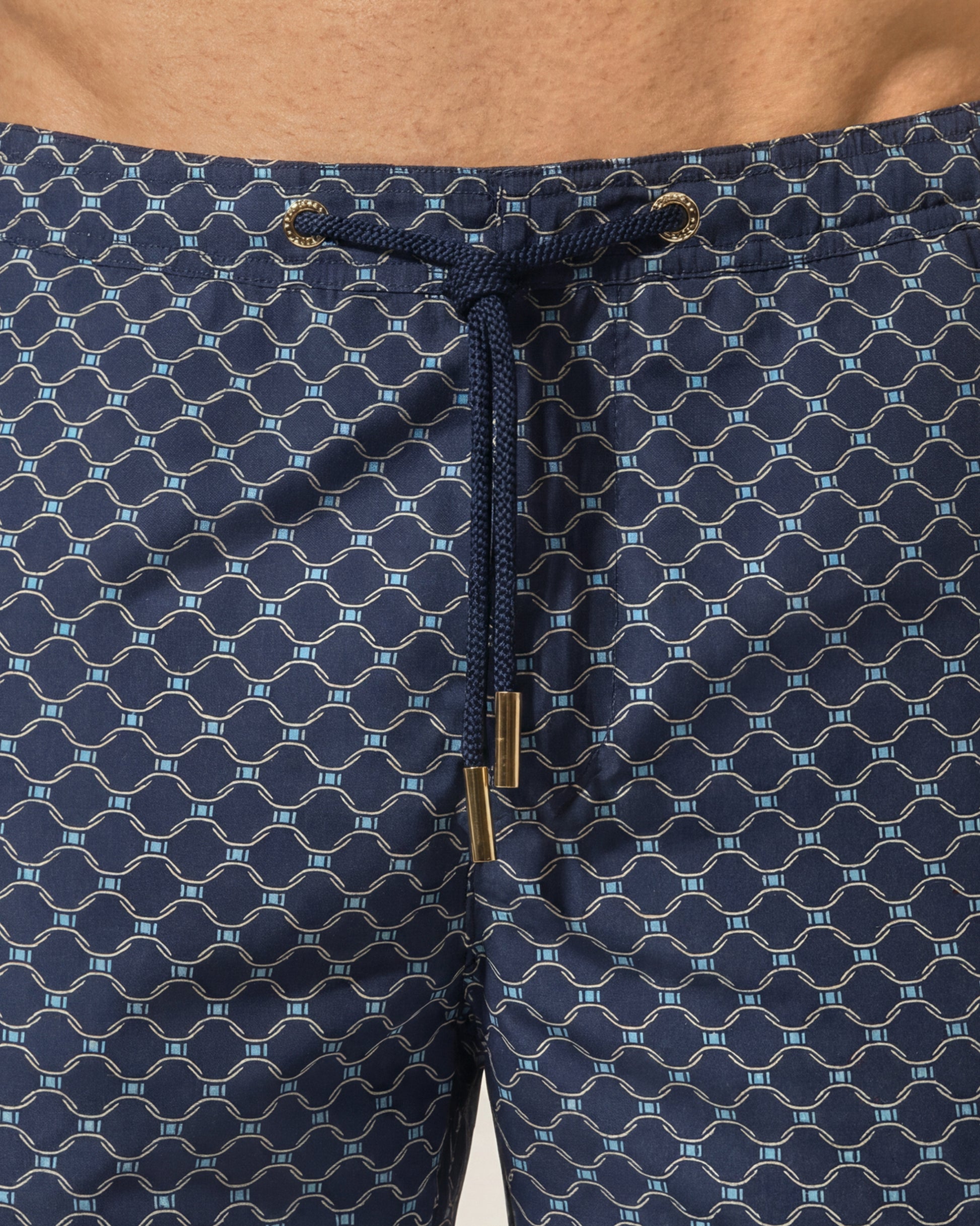 Close-up of a person wearing navy blue patterned swim trunks with a matching drawstring and gold-tone hardware.