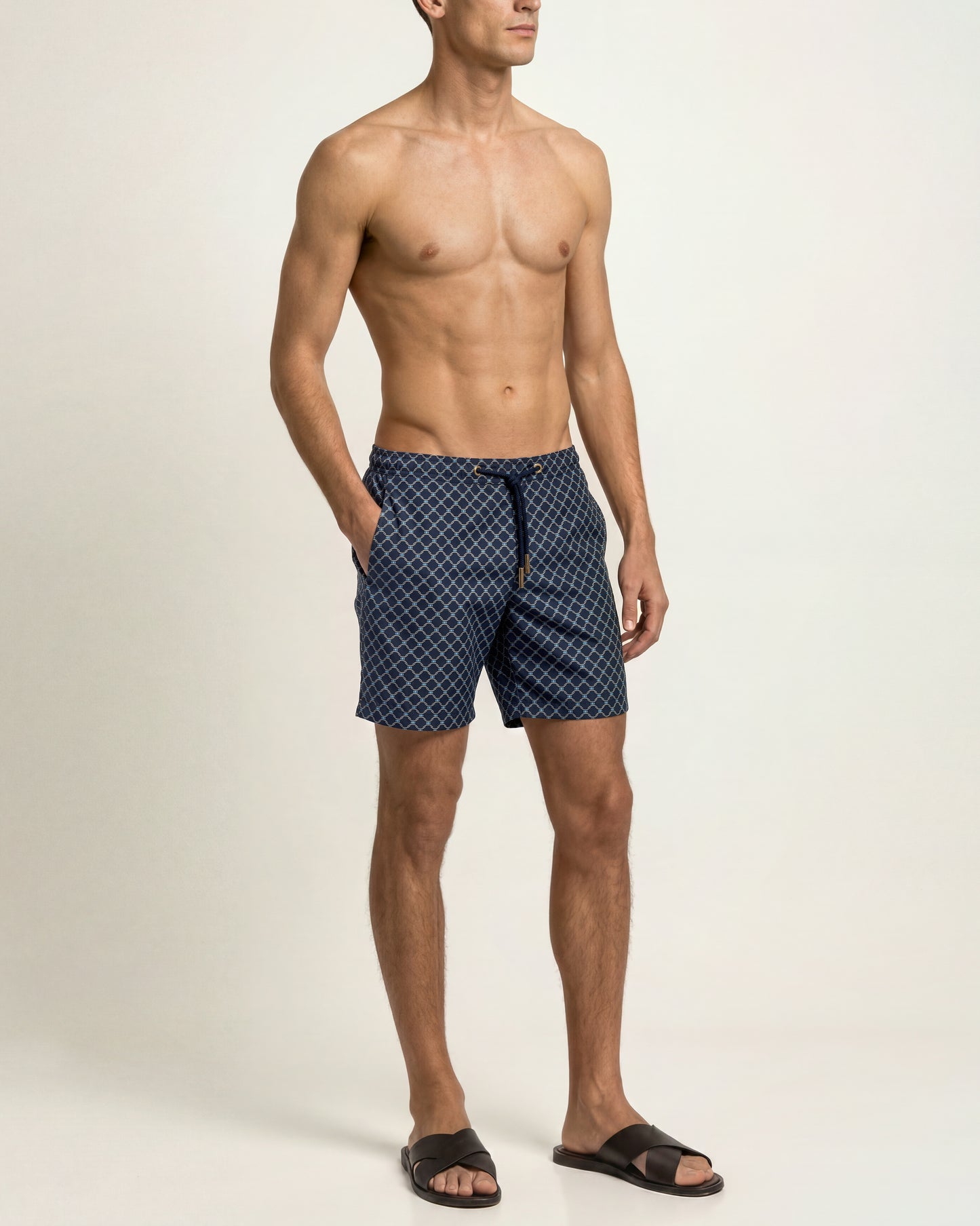 A shirtless male model wearing navy blue patterned swim trunks and black slide sandals, standing against a plain off-white studio background.