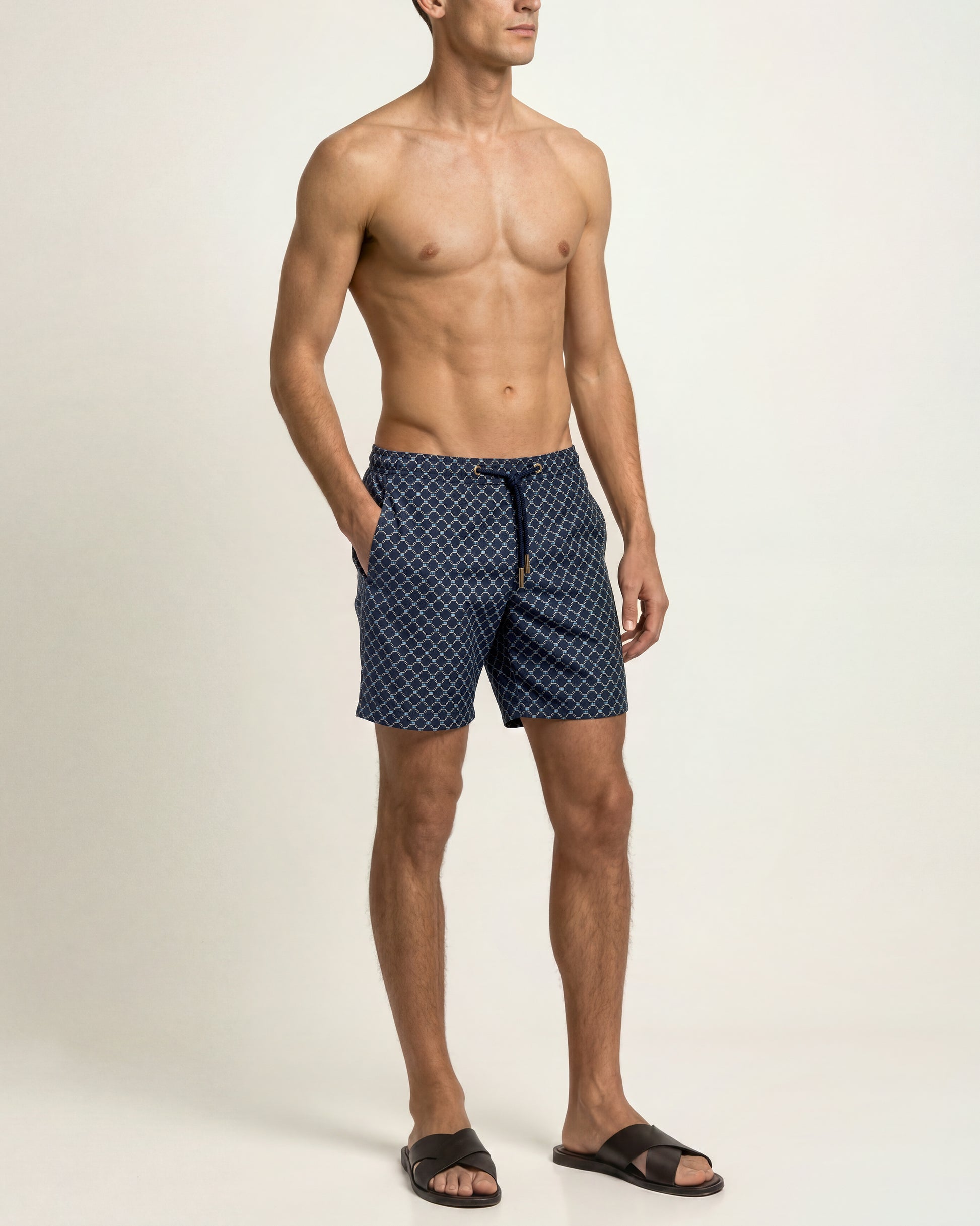 A shirtless male model wearing navy blue patterned swim trunks and black slide sandals, standing against a plain off-white studio background.
