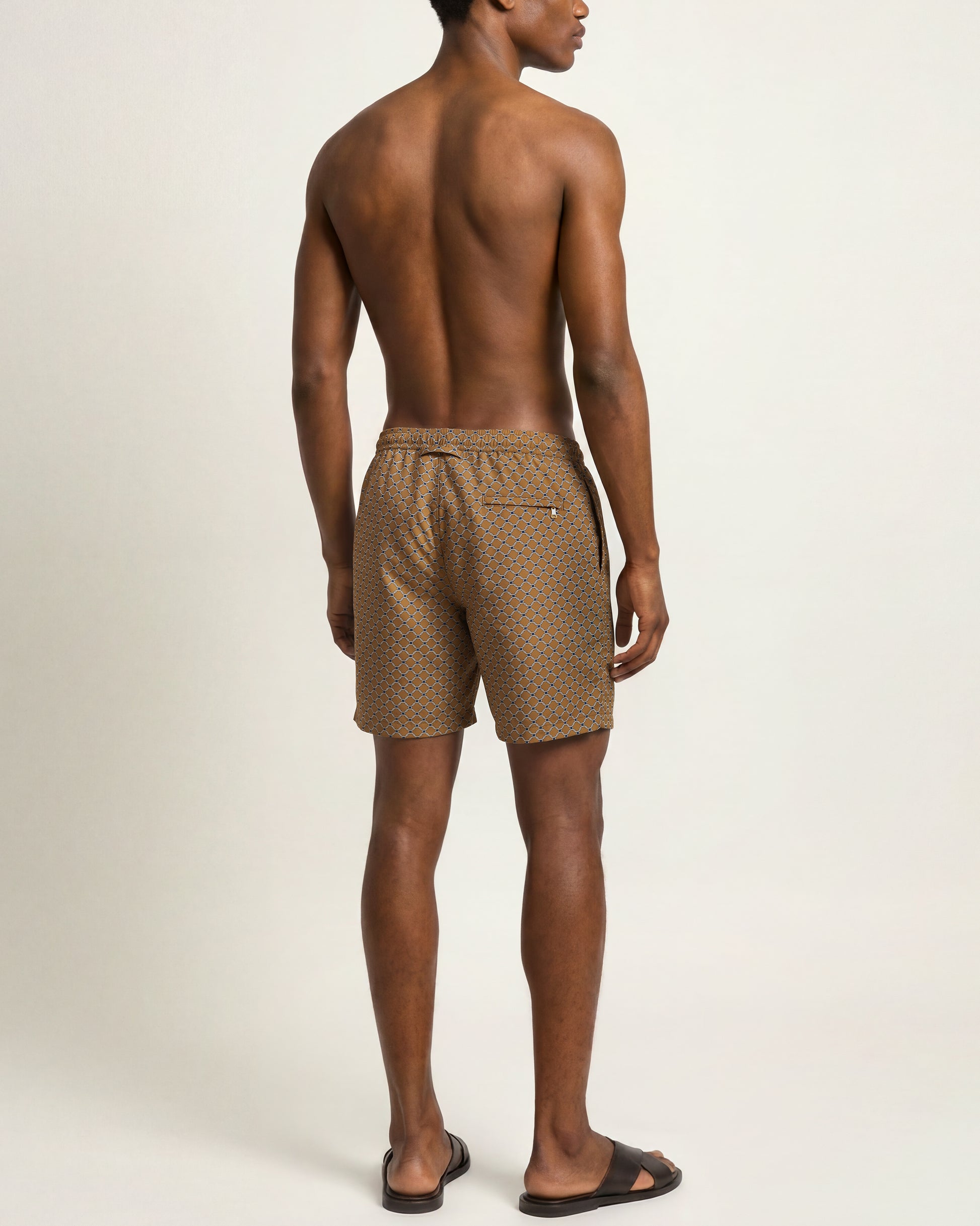 A man shown from the back wearing brown and beige checkered swim trunks and black slide sandals, standing against a plain off-white studio background.