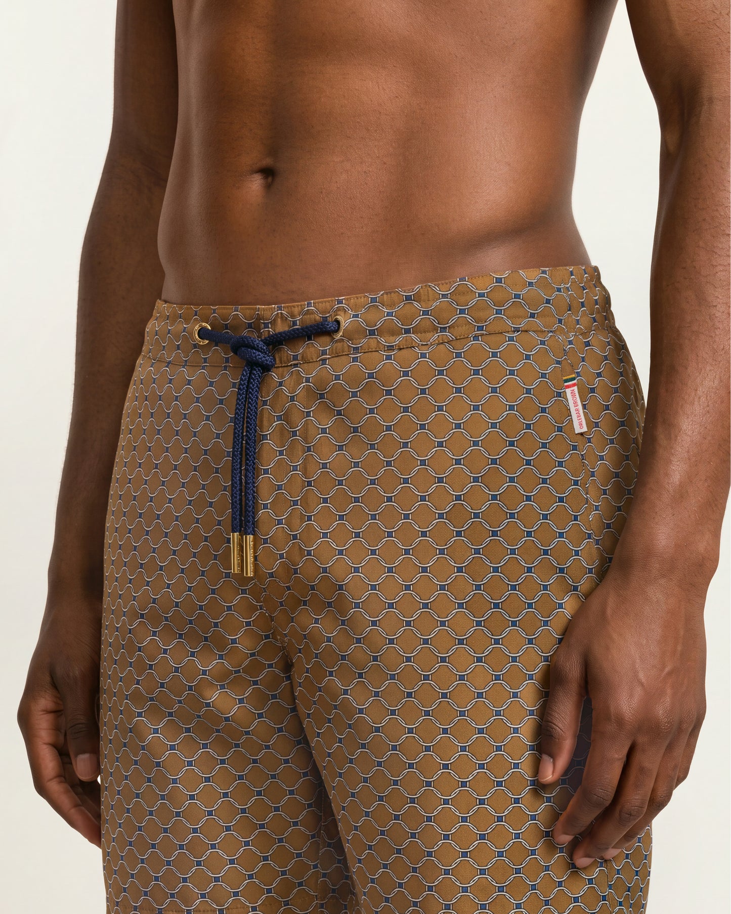 A close-up of a man's lower torso wearing patterned brown drawstring shorts with a geometric lattice design and a navy blue cord.