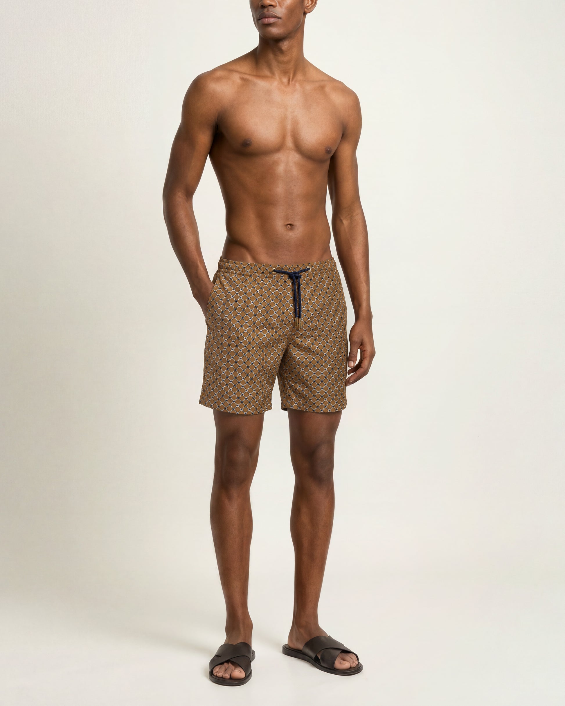 A shirtless male model wearing brown and beige patterned swim trunks with a dark drawstring, standing against a plain off-white studio background.