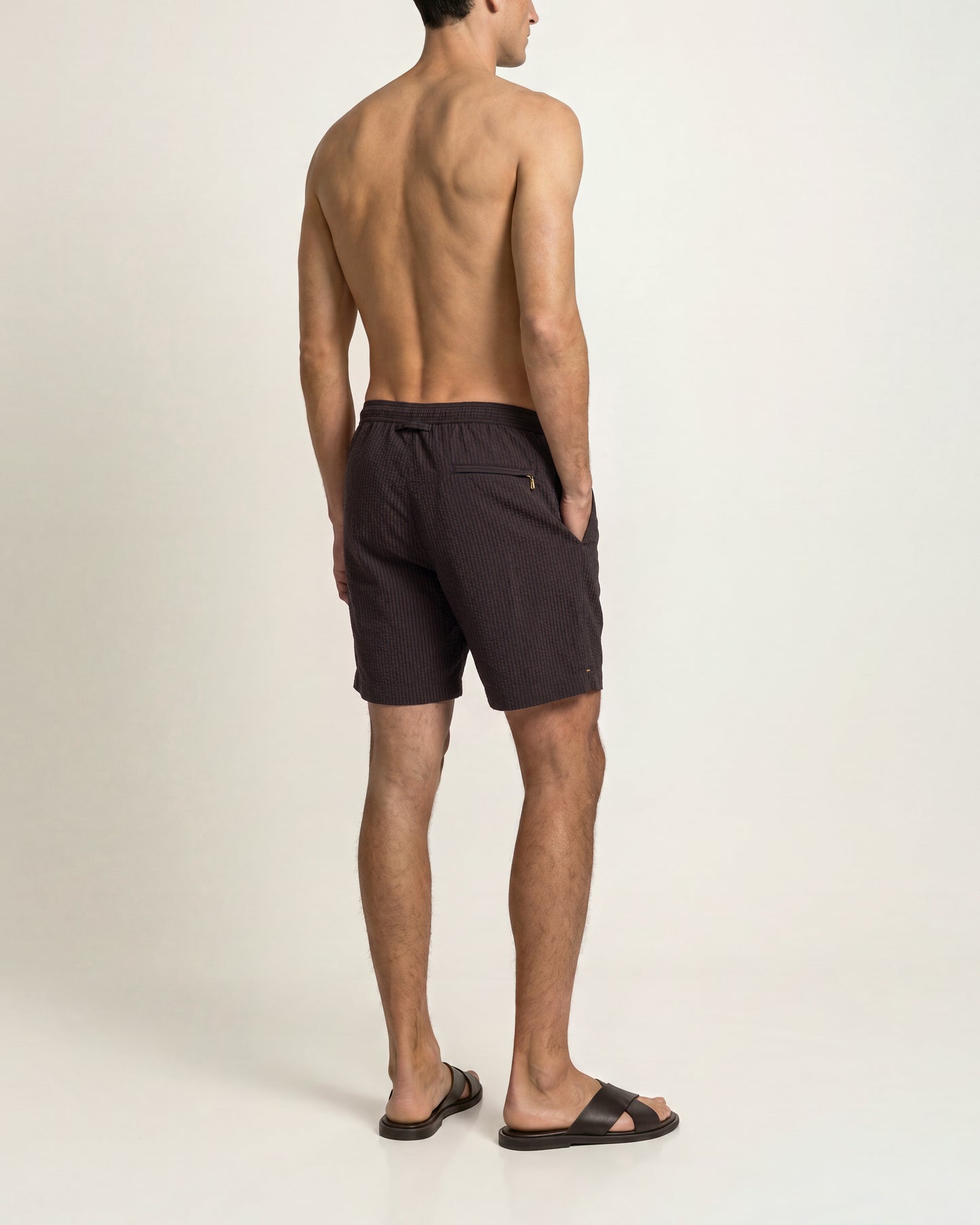A man shown from the back wearing dark brown swim trunks and black slide sandals, standing against a plain off-white studio background.