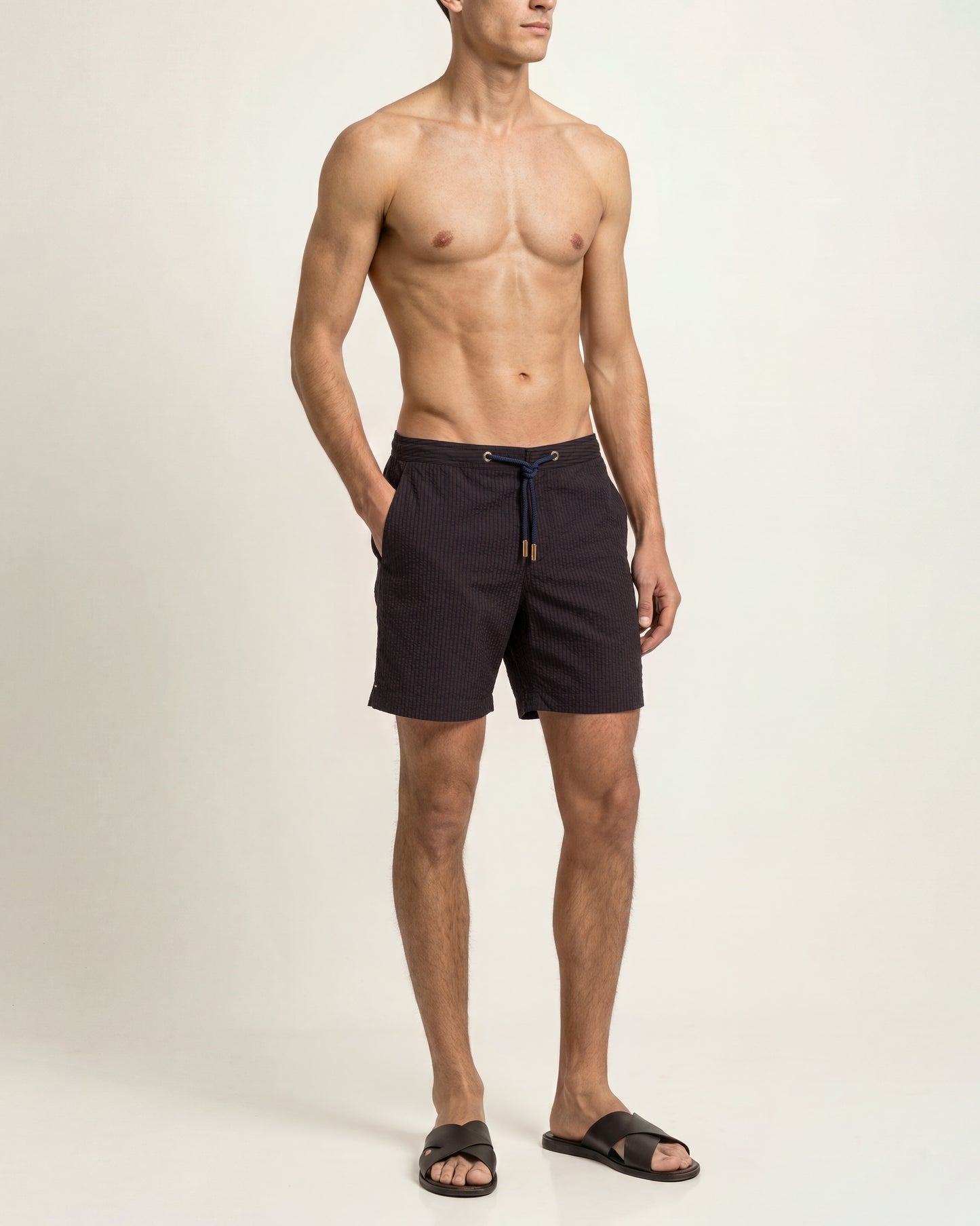 A shirtless male model wearing dark charcoal drawstring swim shorts and black slide sandals, standing against a plain off-white studio background.