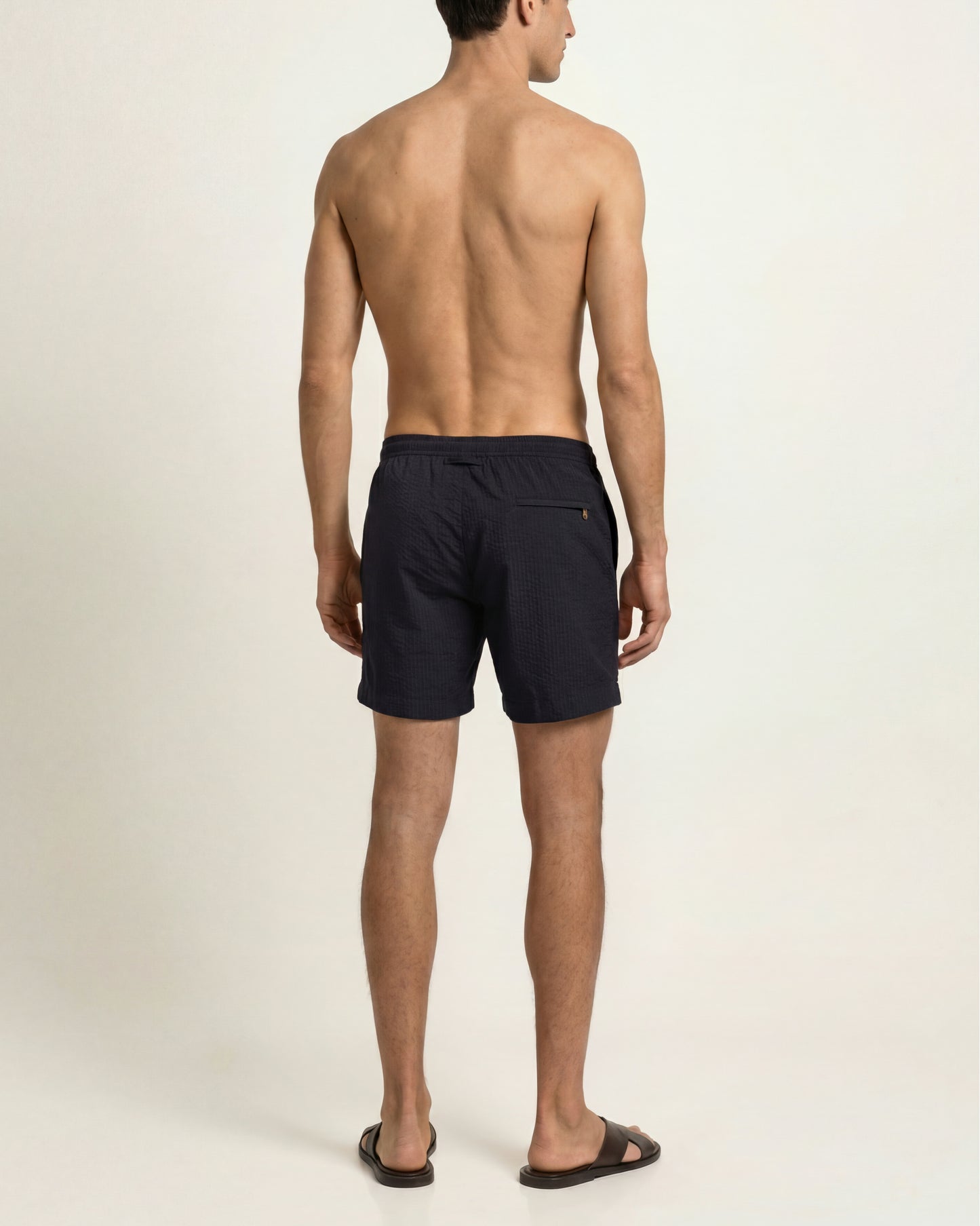 A man shown from the back wearing dark navy swim shorts and brown slide sandals against a plain light beige studio background.