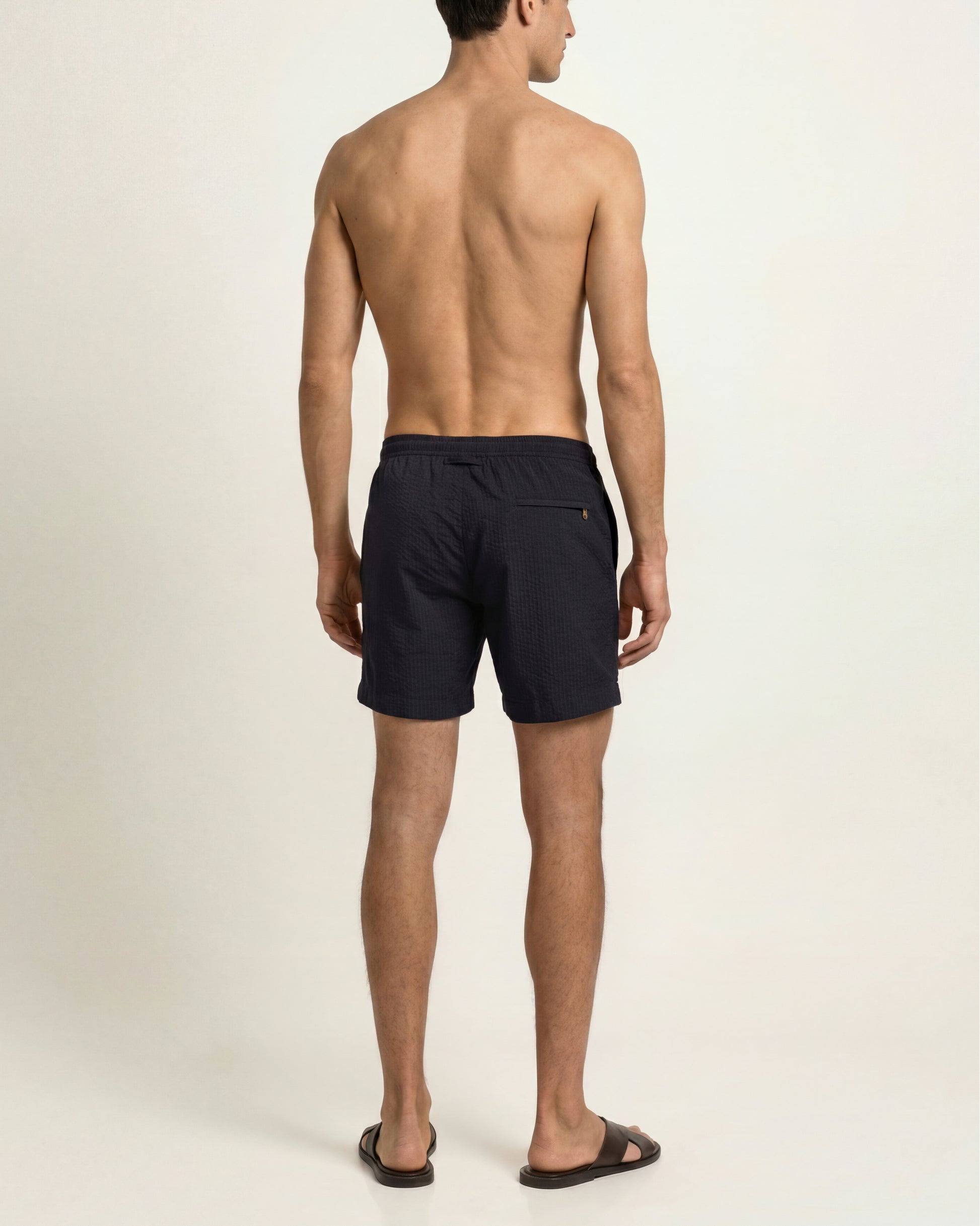 A man shown from the back wearing dark navy swim shorts and brown slide sandals against a plain light beige studio background.