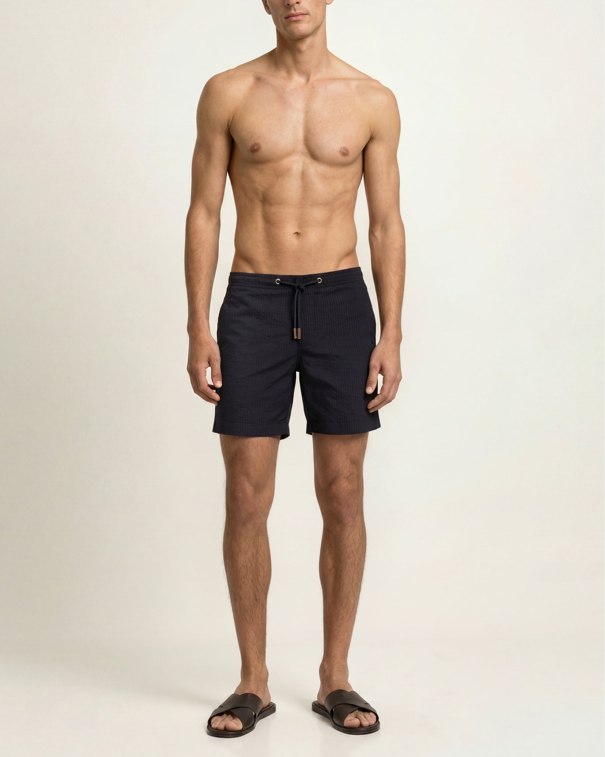 A shirtless male model wearing dark navy drawstring swim shorts and dark brown slide sandals, standing against a plain off-white studio background.