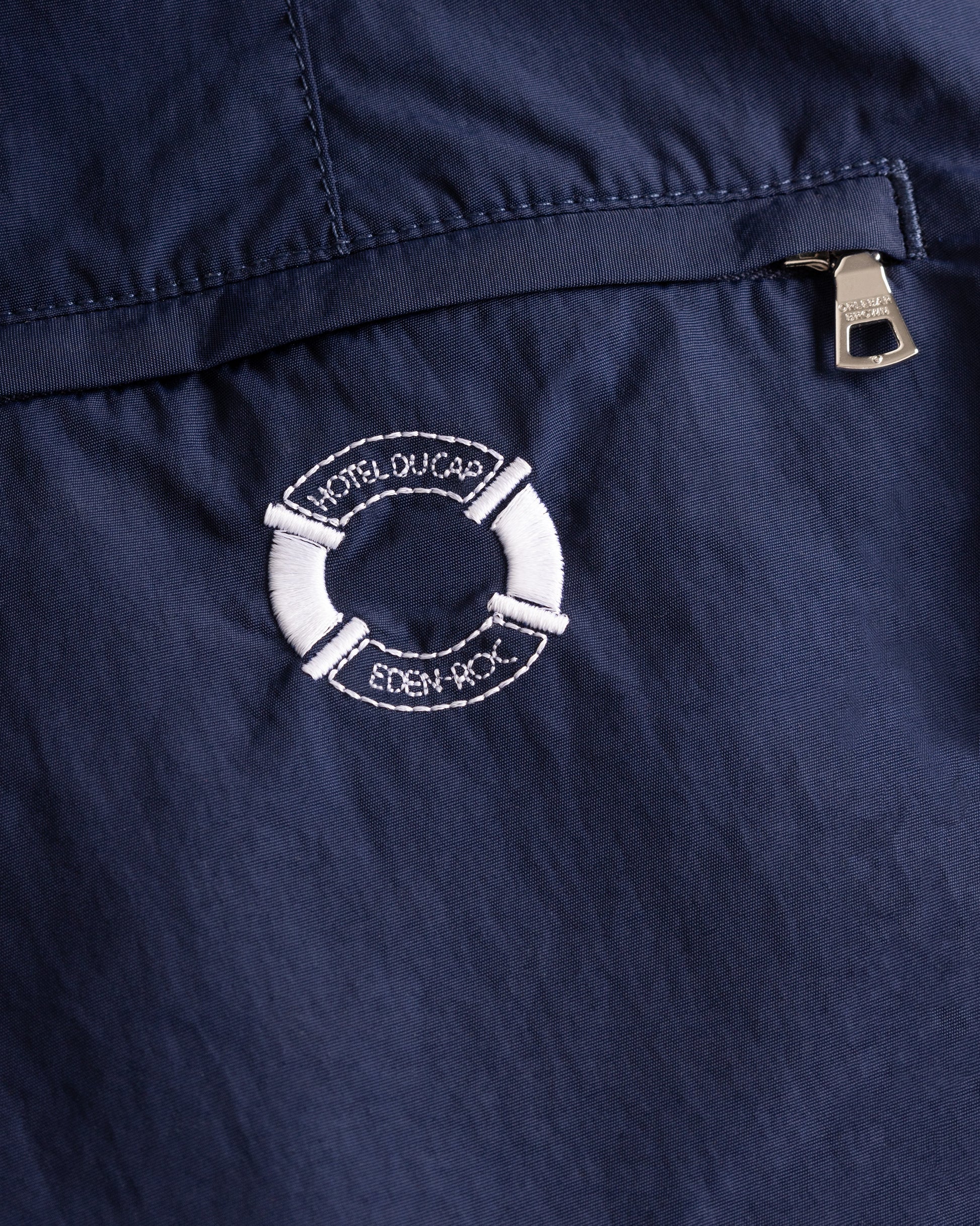 Close-up of a navy blue garment, likely a jacket or coat, featuring a white embroidered life preserver logo with the text 'HOTEL DU CAP' and 'EDEN ROC' on the chest, next to a silver zipper pull.