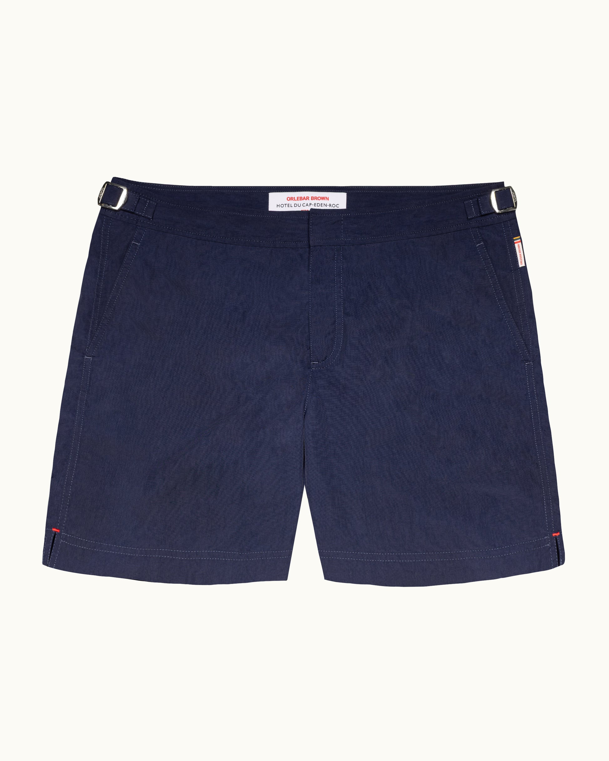 A pair of dark navy blue shorts with a subtle textured pattern, featuring belt loops, side pockets, and a small red and white brand tag on the right side, displayed against a light off-white background.