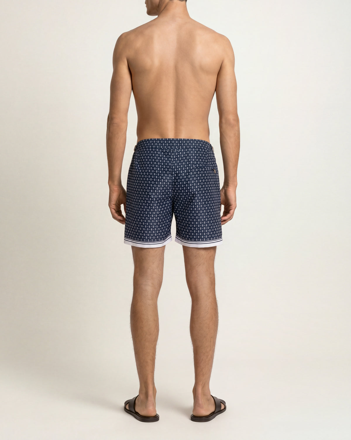 A man viewed from behind wearing navy blue swim trunks with a white geometric pattern and white trim, standing against a white studio background.