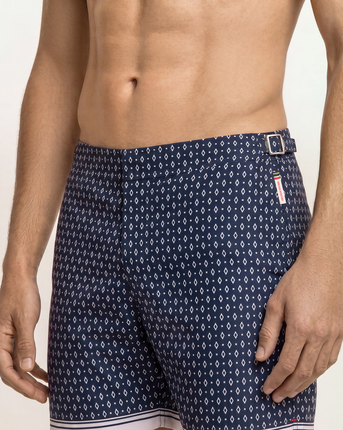 A close-up of a man's torso wearing navy blue swim trunks with a white geometric pattern, featuring a small white and red brand tag on the side.