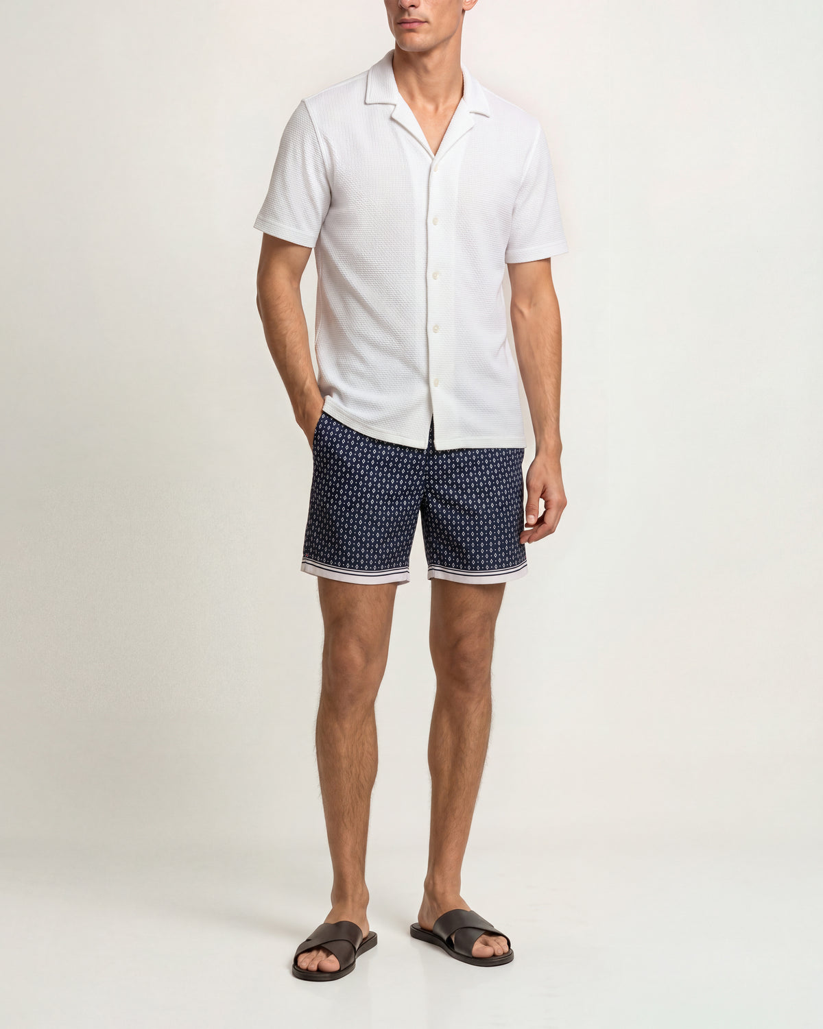 A man modeling a white short-sleeve button-up shirt and navy blue shorts with a white polka-dot pattern, paired with dark brown slide sandals, against a plain off-white studio background.