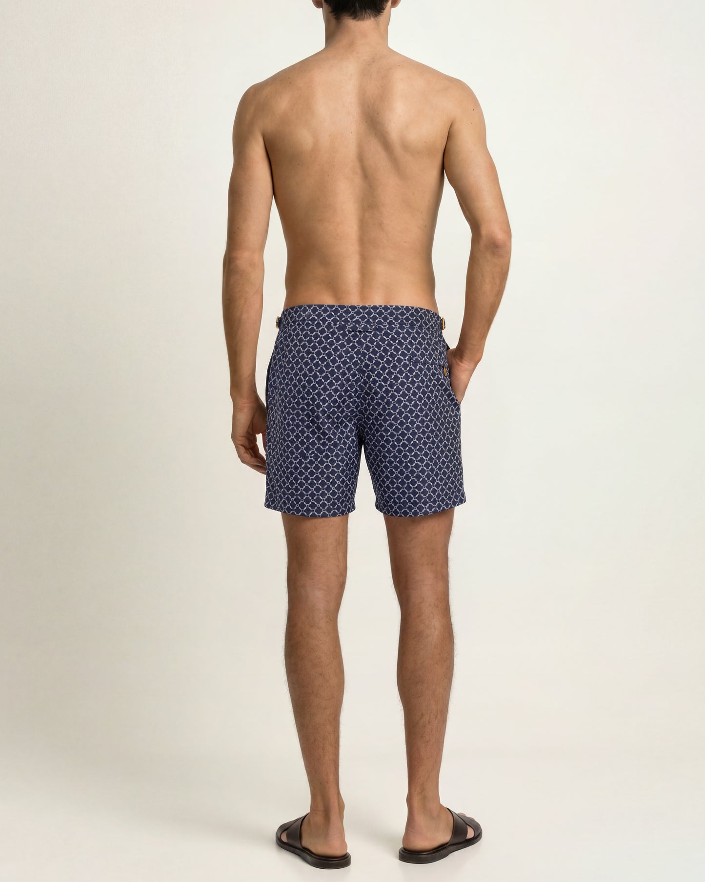 A man viewed from the back wearing navy blue swim trunks with a small white geometric pattern, standing against a plain light beige studio background.
