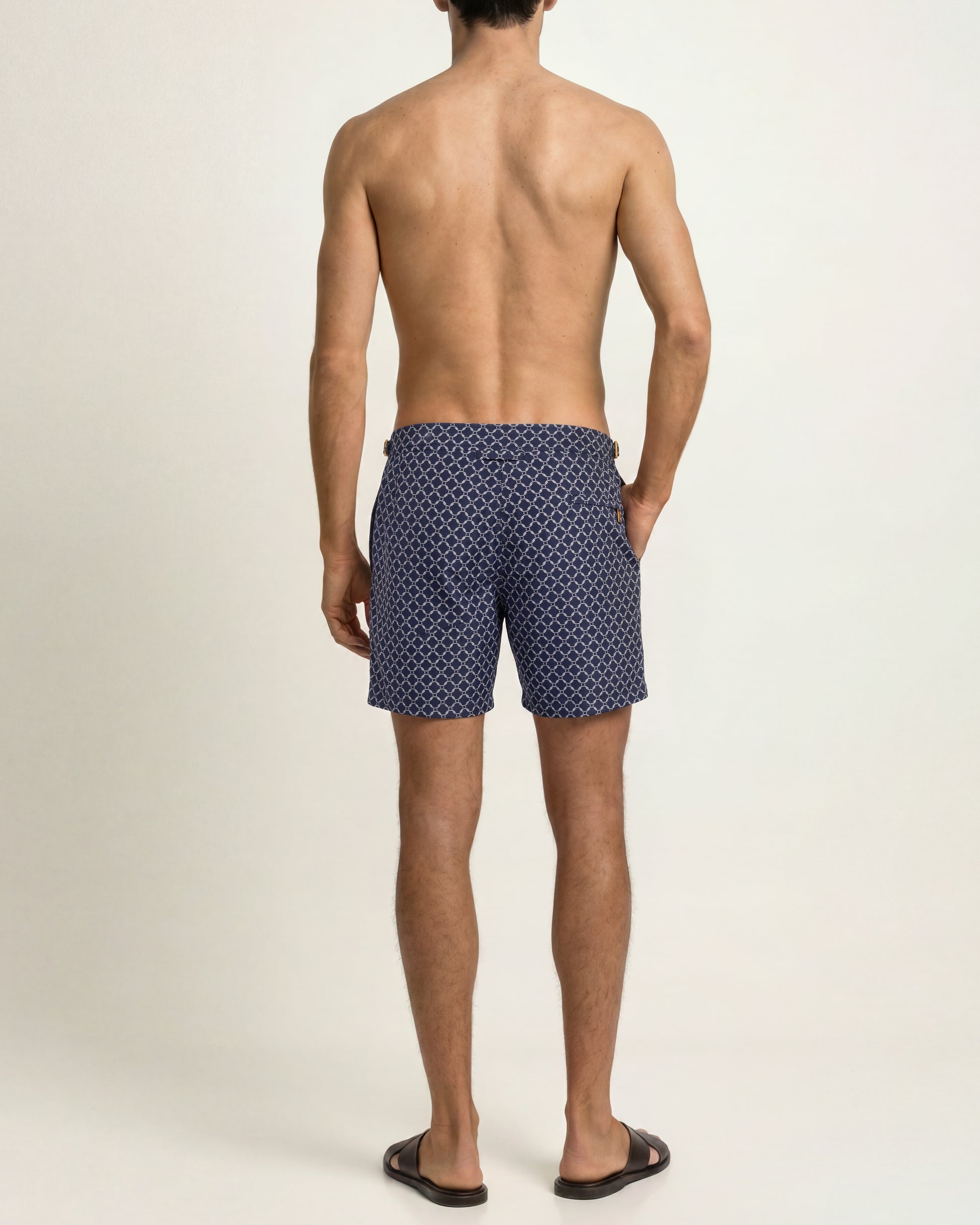 A man viewed from the back wearing navy blue swim trunks with a small white geometric pattern, standing against a plain light beige studio background.