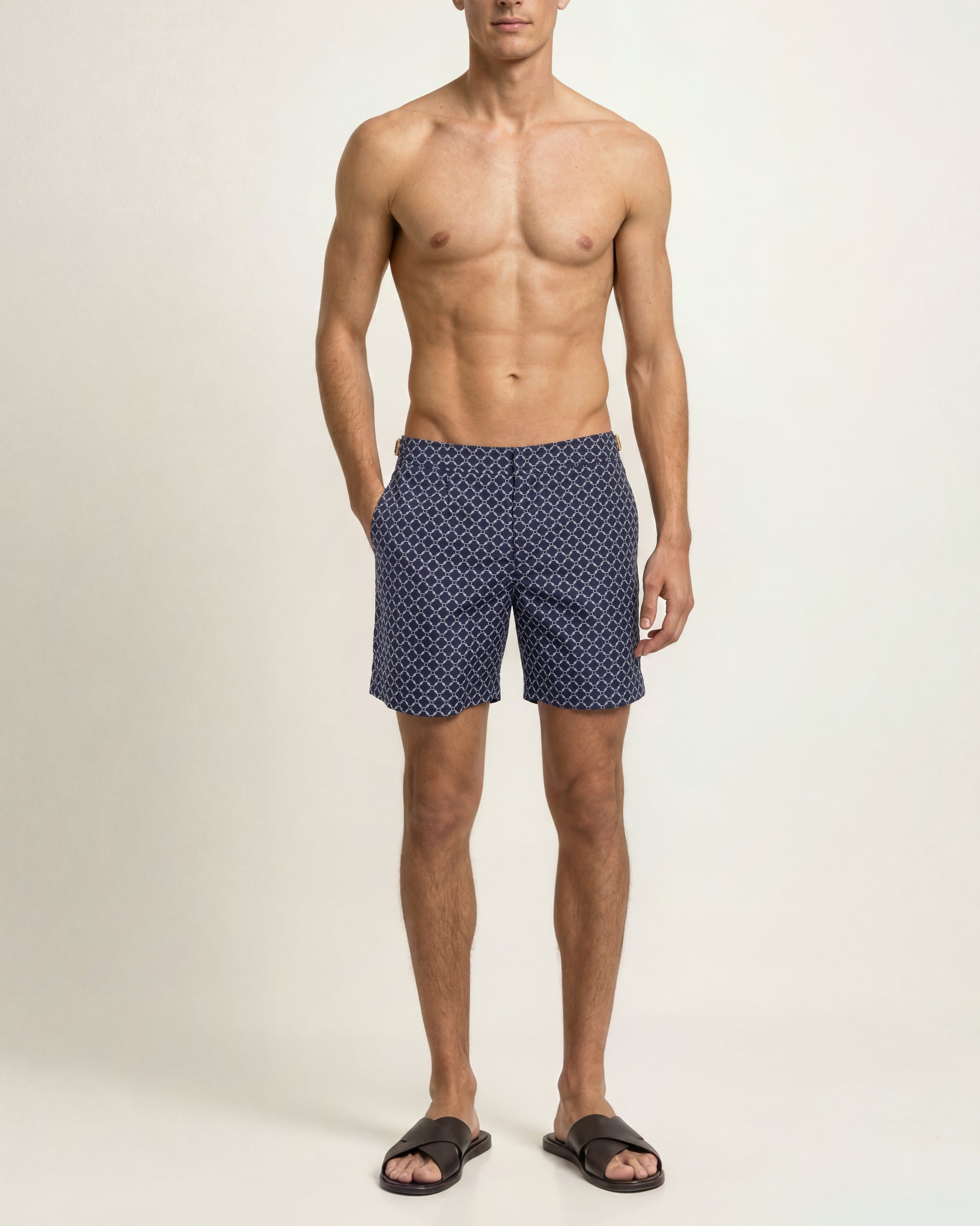 A shirtless male model wearing navy blue swim trunks with a small white geometric pattern, standing against a plain off-white studio background.