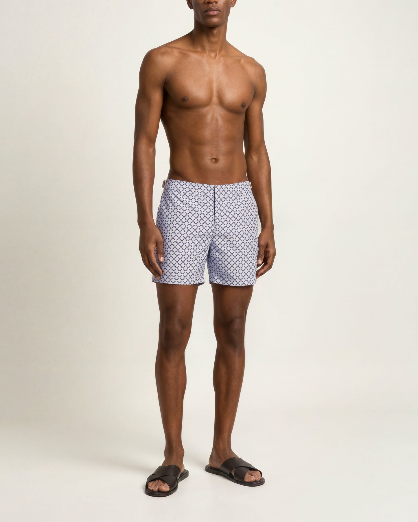 A shirtless male model wearing blue and white patterned swim trunks and dark brown slide sandals, standing against a plain off-white studio background.