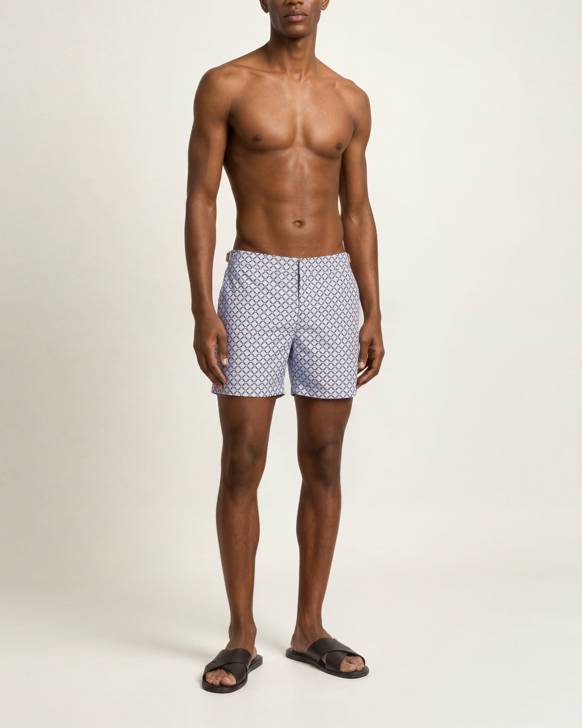 A shirtless male model wearing blue and white patterned swim trunks and dark brown slide sandals, standing against a plain off-white studio background.