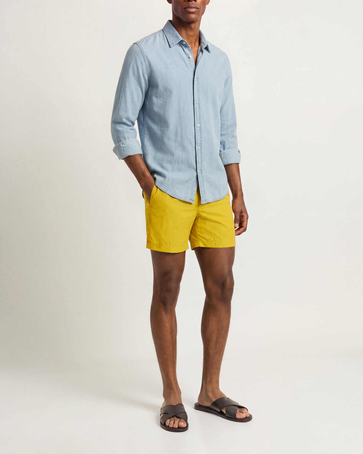 A man modeling a light blue long-sleeve button-up shirt and bright yellow shorts, paired with dark brown slide sandals, against a plain off-white studio background.