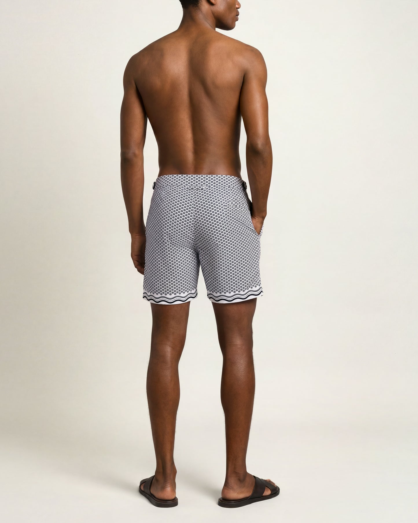 A man shown from the back wearing patterned swim trunks with a black and white geometric print and wavy trim, standing against a plain light beige studio background.
