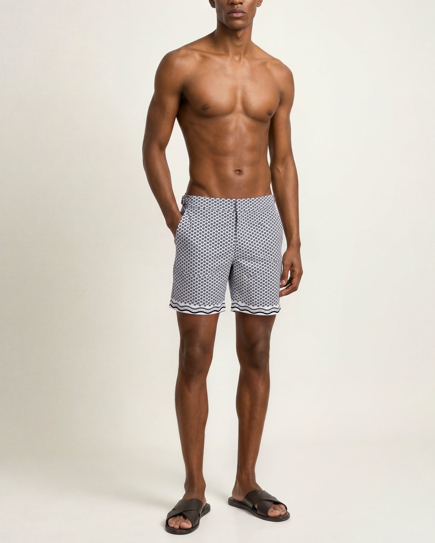 A man modeling patterned swim trunks with a black and white geometric print and wavy trim, standing against a plain light beige studio background.