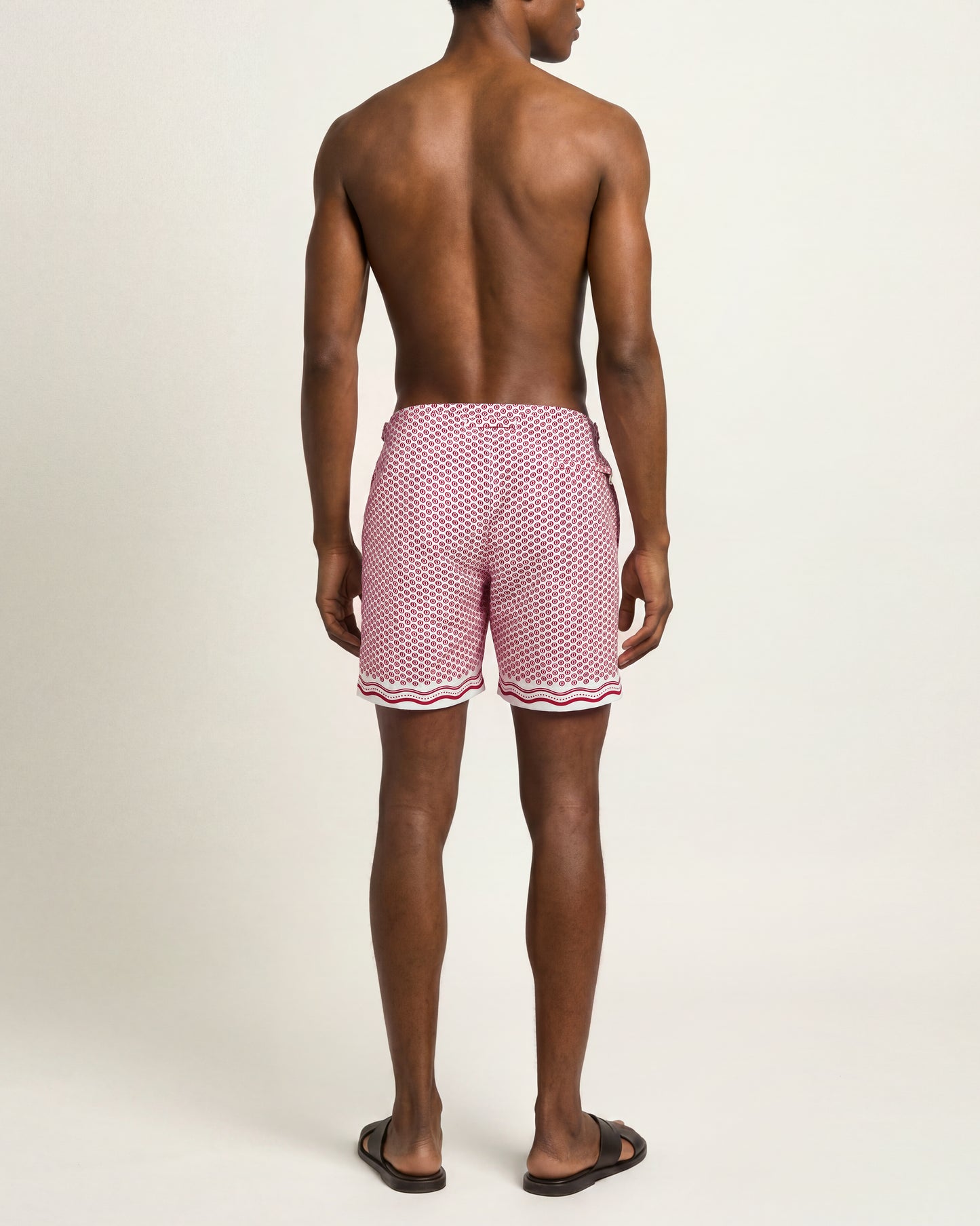 A man viewed from the back wearing pink swim trunks with a white geometric pattern and a wavy white stripe at the hem, standing against a plain light beige studio background.