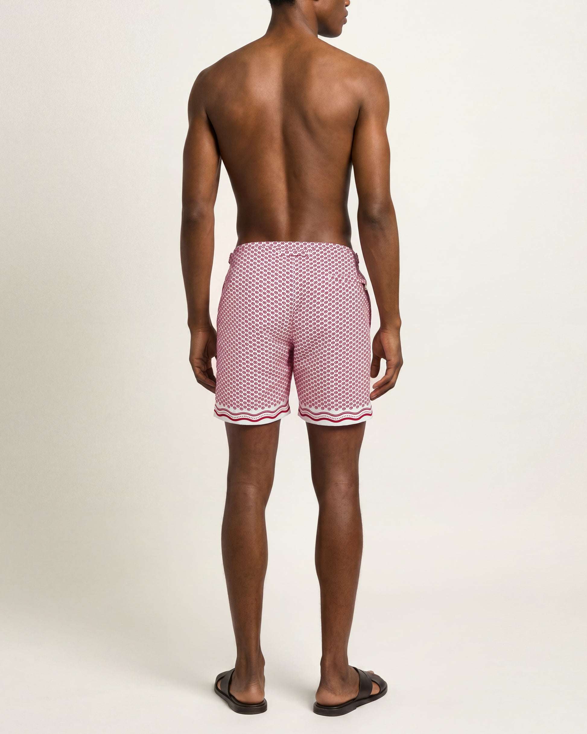 A man viewed from the back wearing pink swim trunks with a white geometric pattern and a wavy white stripe at the hem, standing against a plain light beige studio background.
