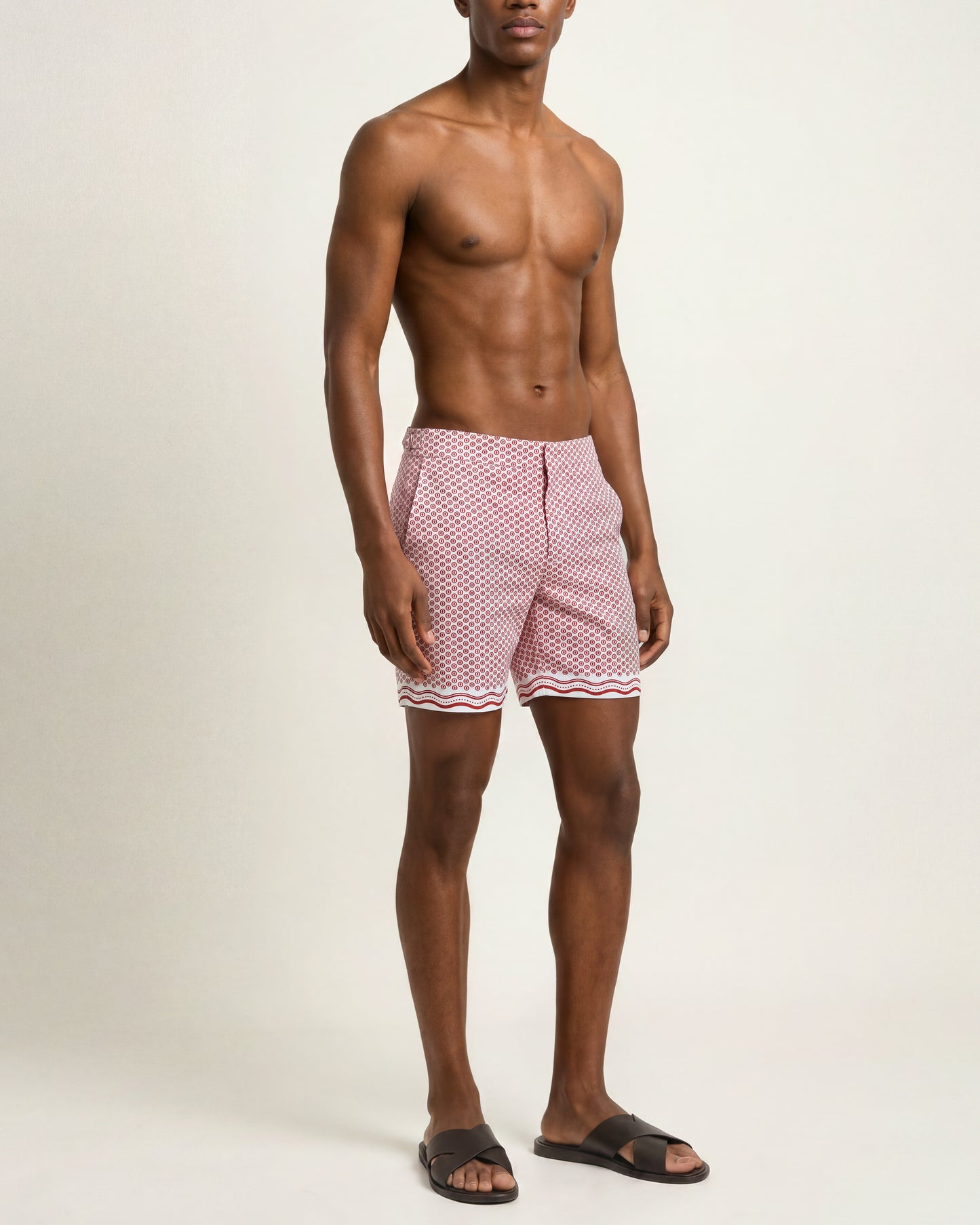 A shirtless male model wearing pink and white patterned swim trunks with a wavy trim, standing against a plain light beige studio background.