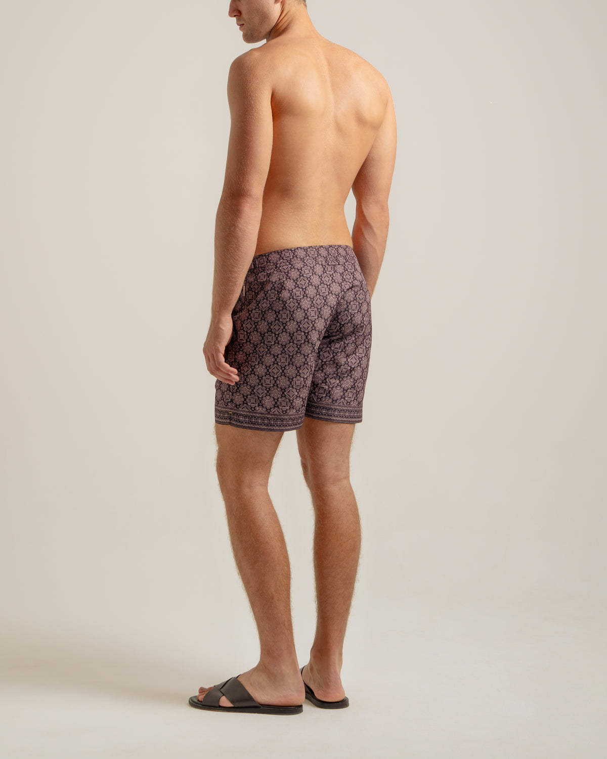A man shown from the back wearing patterned dark brown and purple swim trunks, standing against a plain light beige studio background.