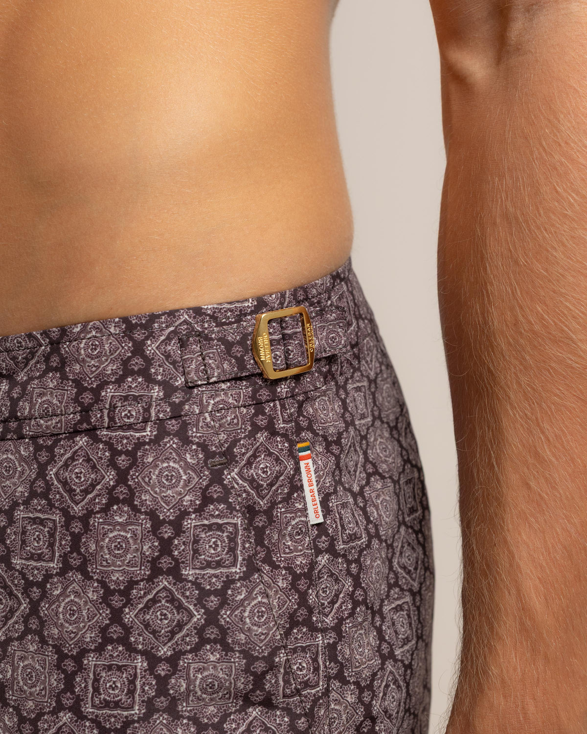 Close-up of a person wearing patterned trousers with a geometric medallion print in dark gray and white, featuring a gold-tone belt buckle and a small red, white, and blue brand tag.