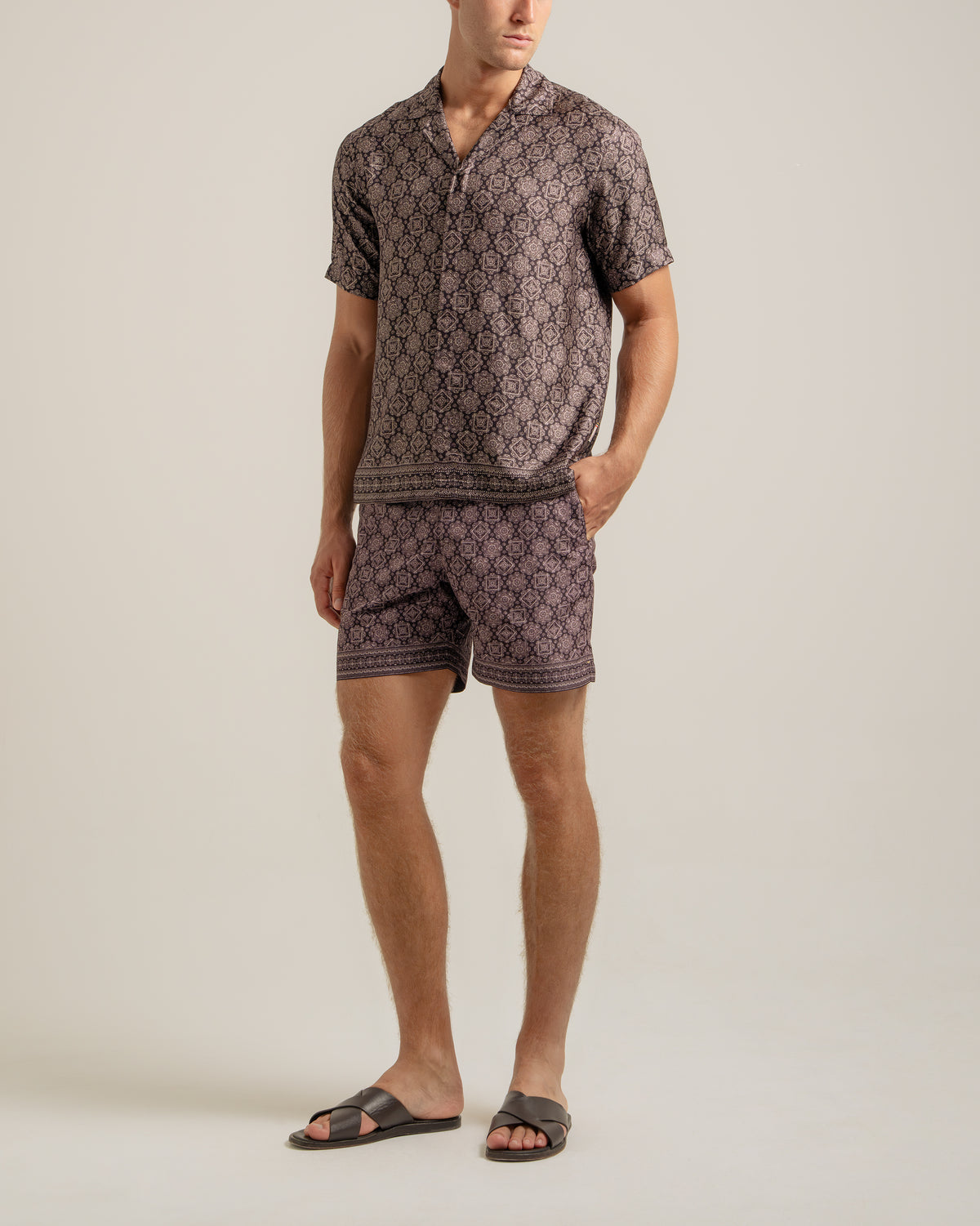 A man modeling a matching short-sleeve button-front shirt and shorts set with an allover geometric pattern in muted purple and beige, paired with dark slide sandals, against a plain light beige studio background.