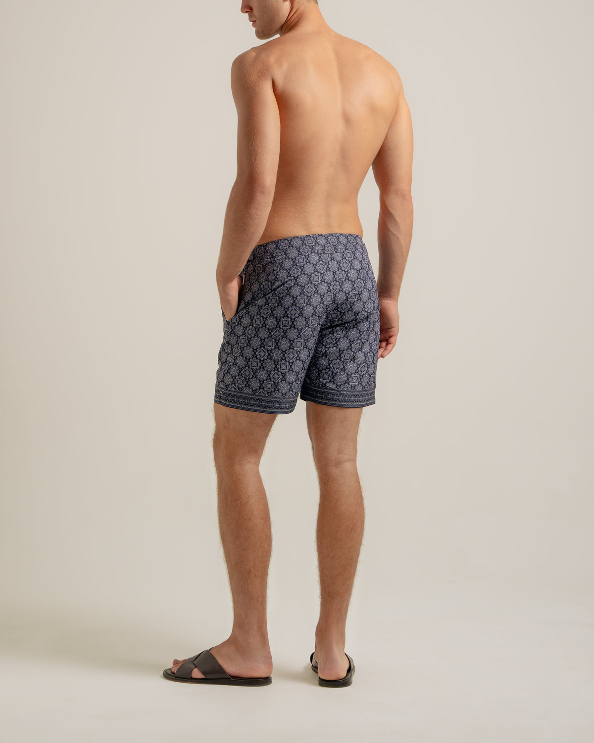 A man shown from the back wearing patterned navy and gray swim trunks and black slide sandals, standing against a plain light beige studio background.
