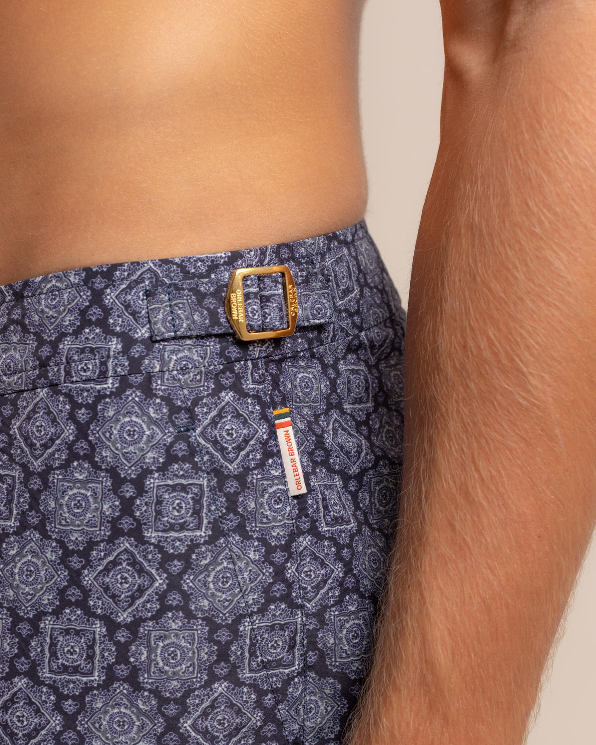 Close-up of a person wearing patterned navy and blue shorts with a gold buckle and a small colorful brand tag.