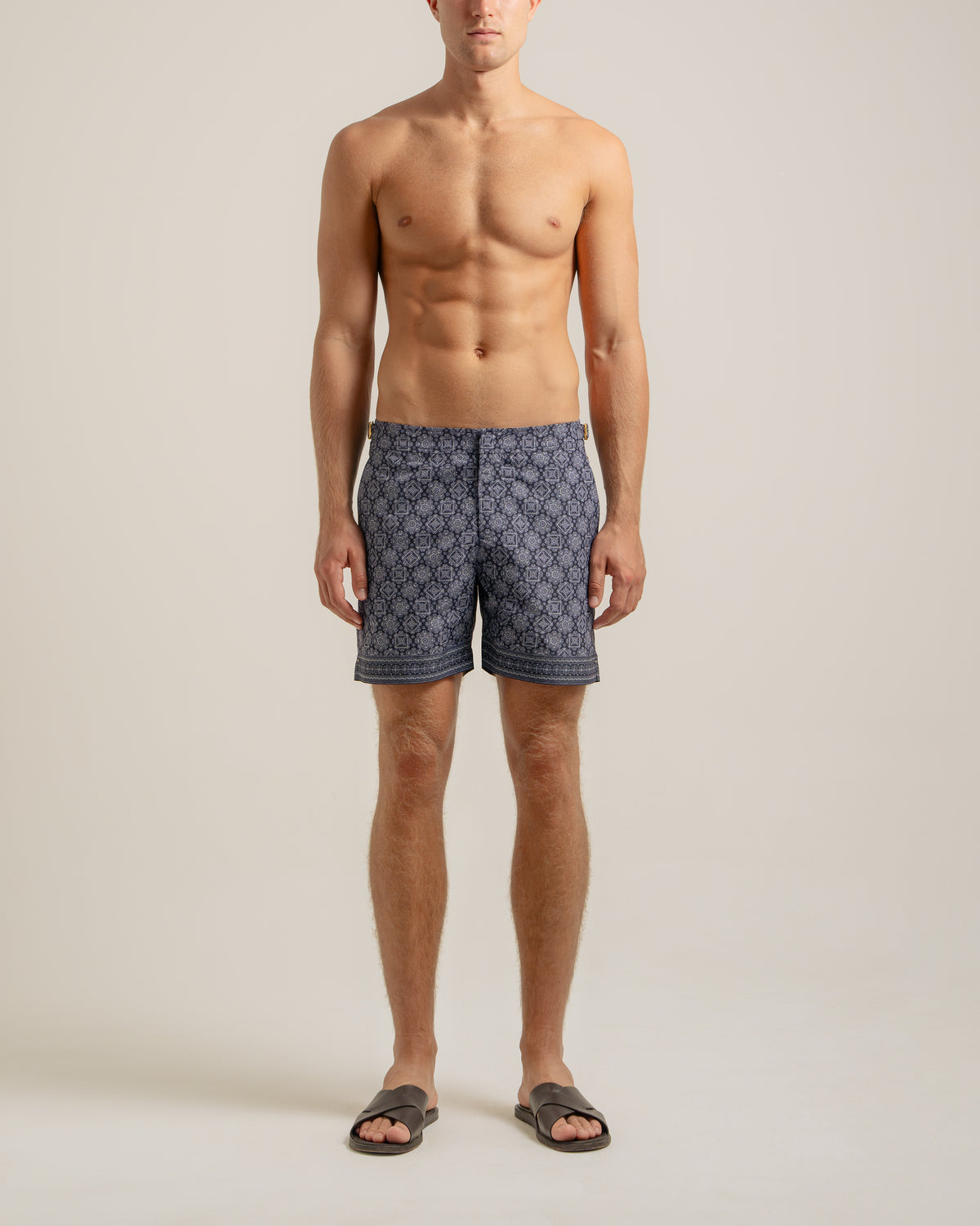 A male model stands against a plain light beige studio background wearing patterned navy and gray swim trunks and dark slide sandals.