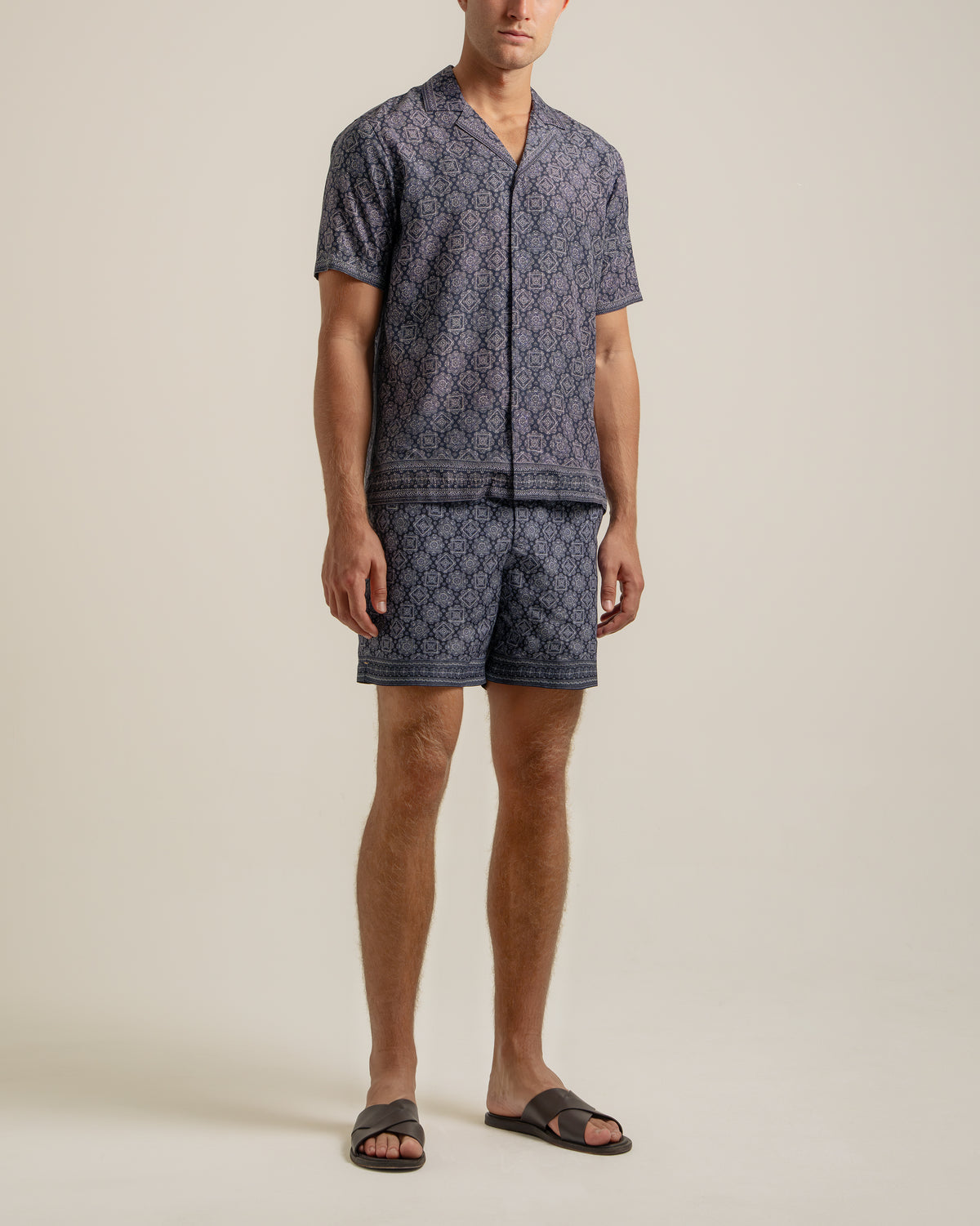 A man modeling a matching short-sleeve button-front pajama top and shorts set in a dark navy blue and light beige geometric pattern, paired with black slide sandals, against a solid light beige studio background.