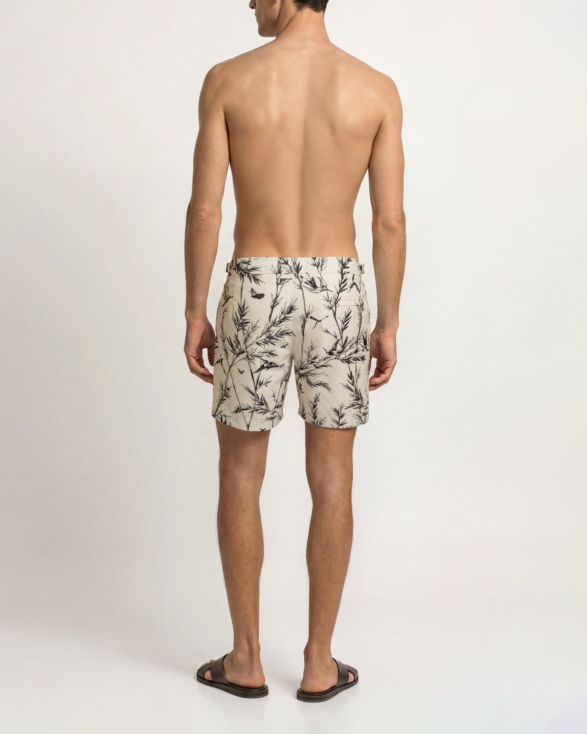 A man viewed from behind wearing beige swim trunks with a black botanical branch pattern, standing against a plain white studio background.