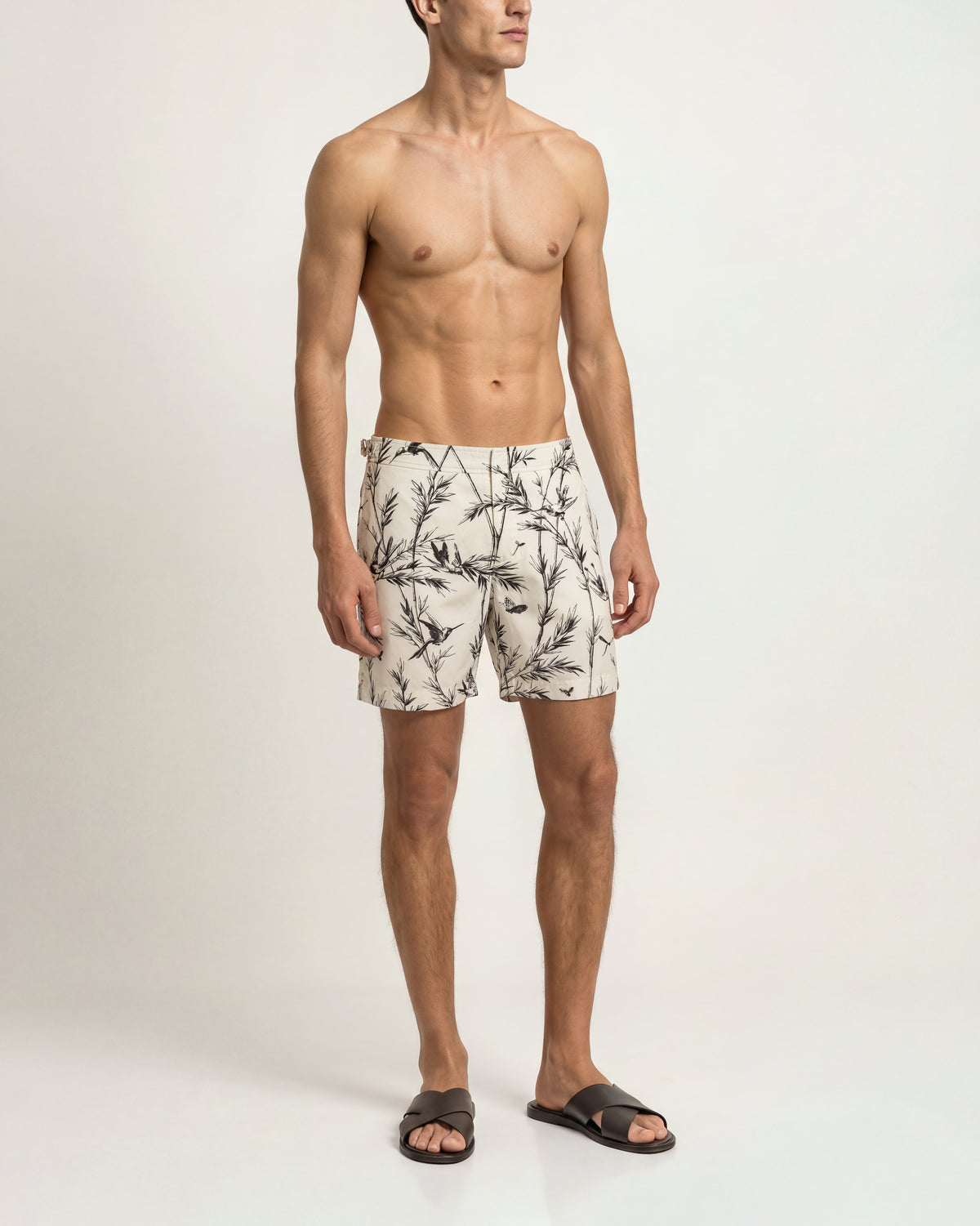 A male model standing against a plain light background, wearing patterned swim trunks with a black tropical leaf print on a cream base, paired with dark slide sandals.