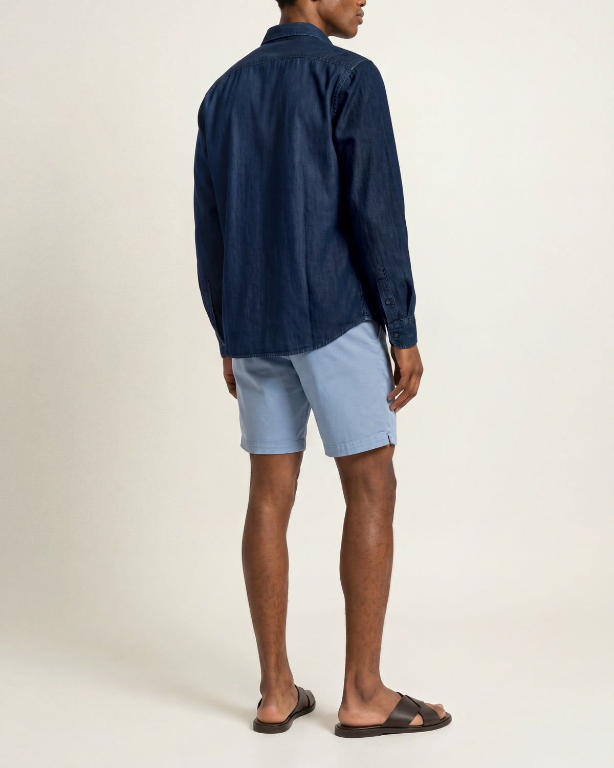 Man wearing light blue shorts with dark denim shirt and brown sandals on a plain background
