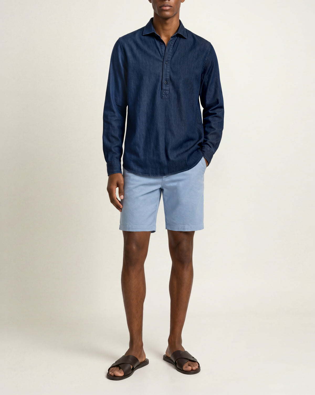 Man wearing light blue shorts with dark denim shirt and brown sandals on a plain background